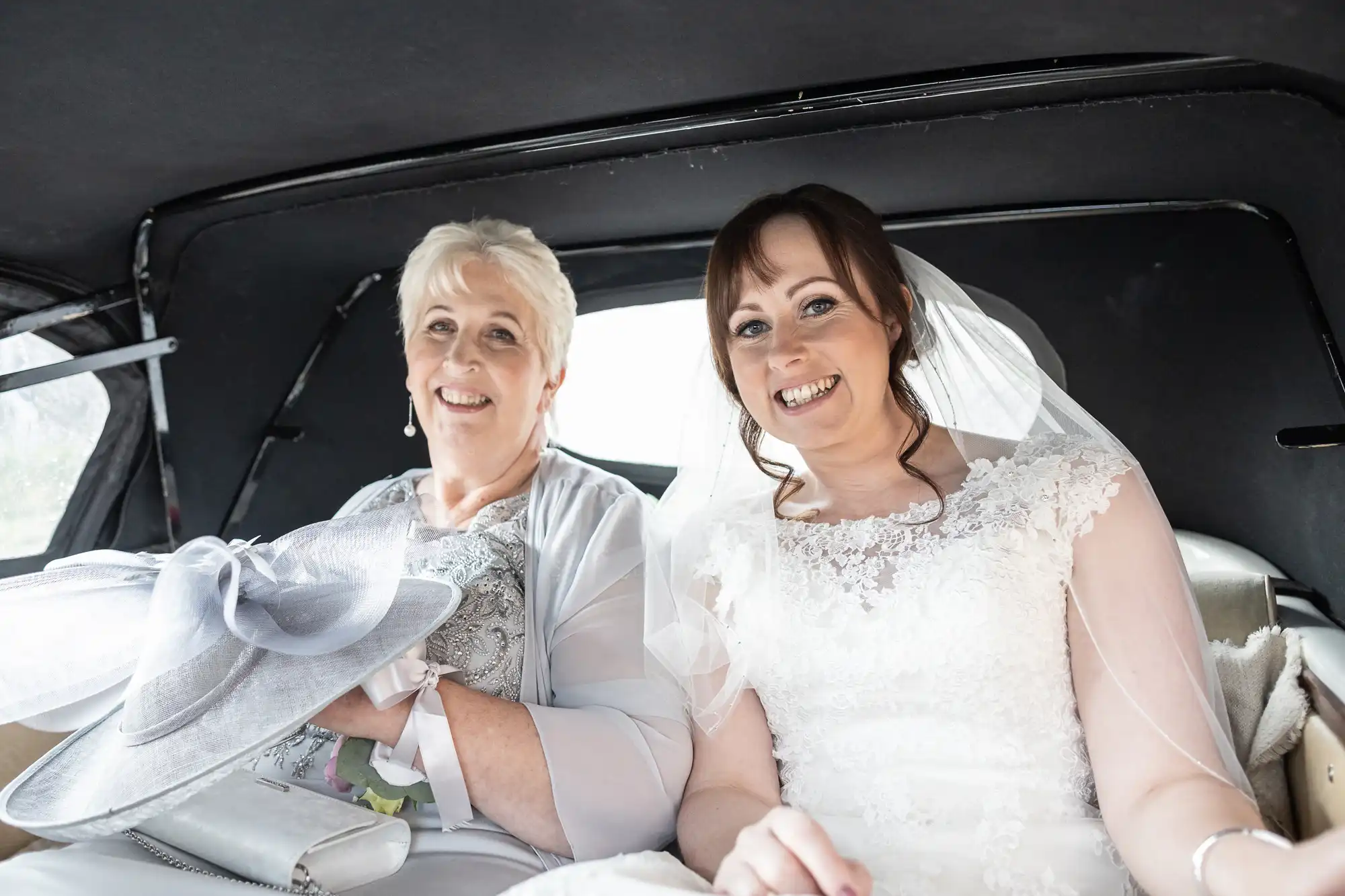 A bride in a white lace wedding dress and veil sits next to an older woman in a light-coloured outfit holding a large hat. Both are smiling happily inside a car, ready for the wedding ceremony, with sunlight coming through the windows.