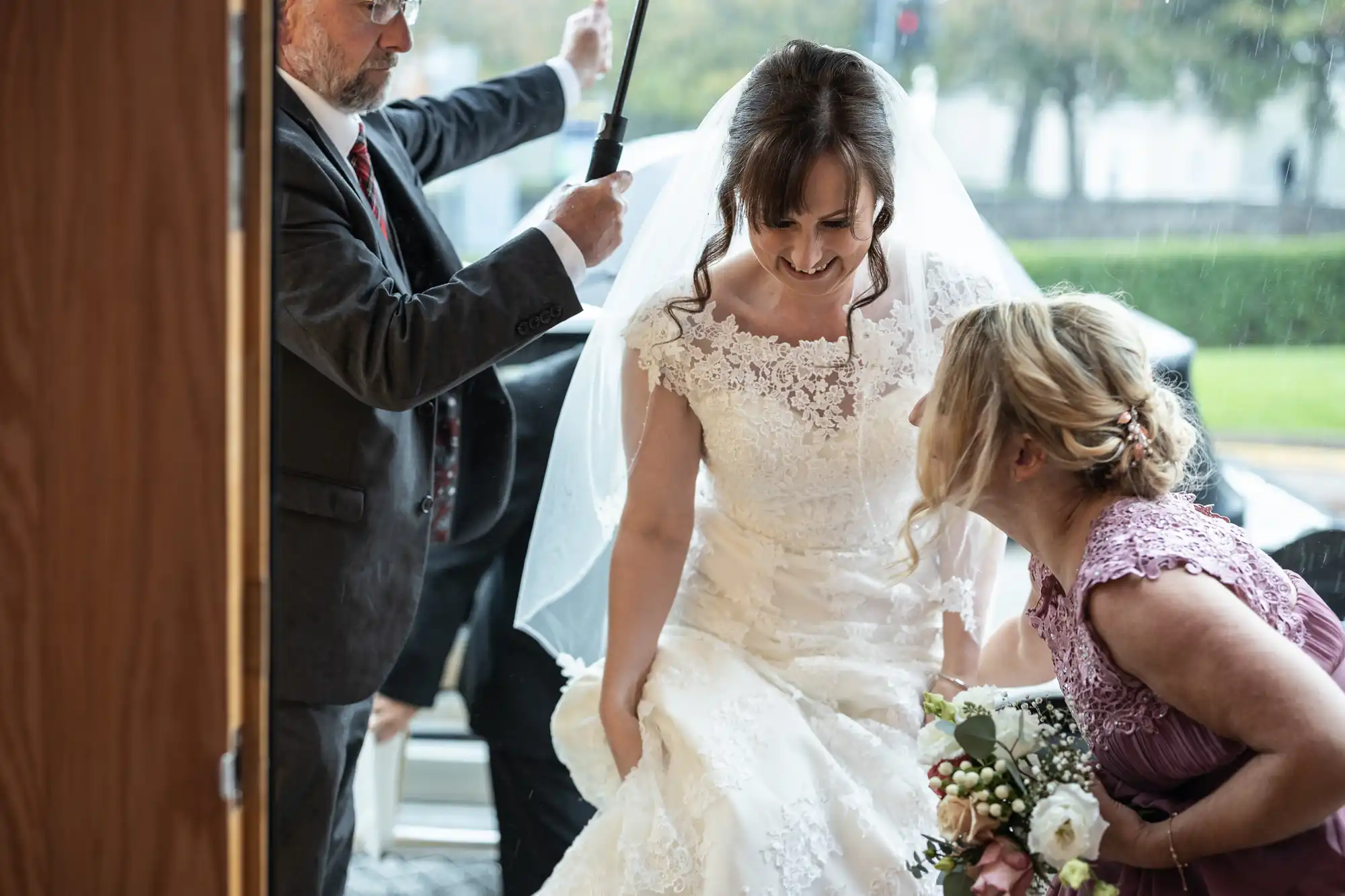 A bride in a lace wedding dress smiles as she steps indoors from the rain. A man holds an umbrella above her and a bridesmaid in a lavender dress kneels, holding a bouquet and arranging the bride's gown. Green grass and trees are visible outside.