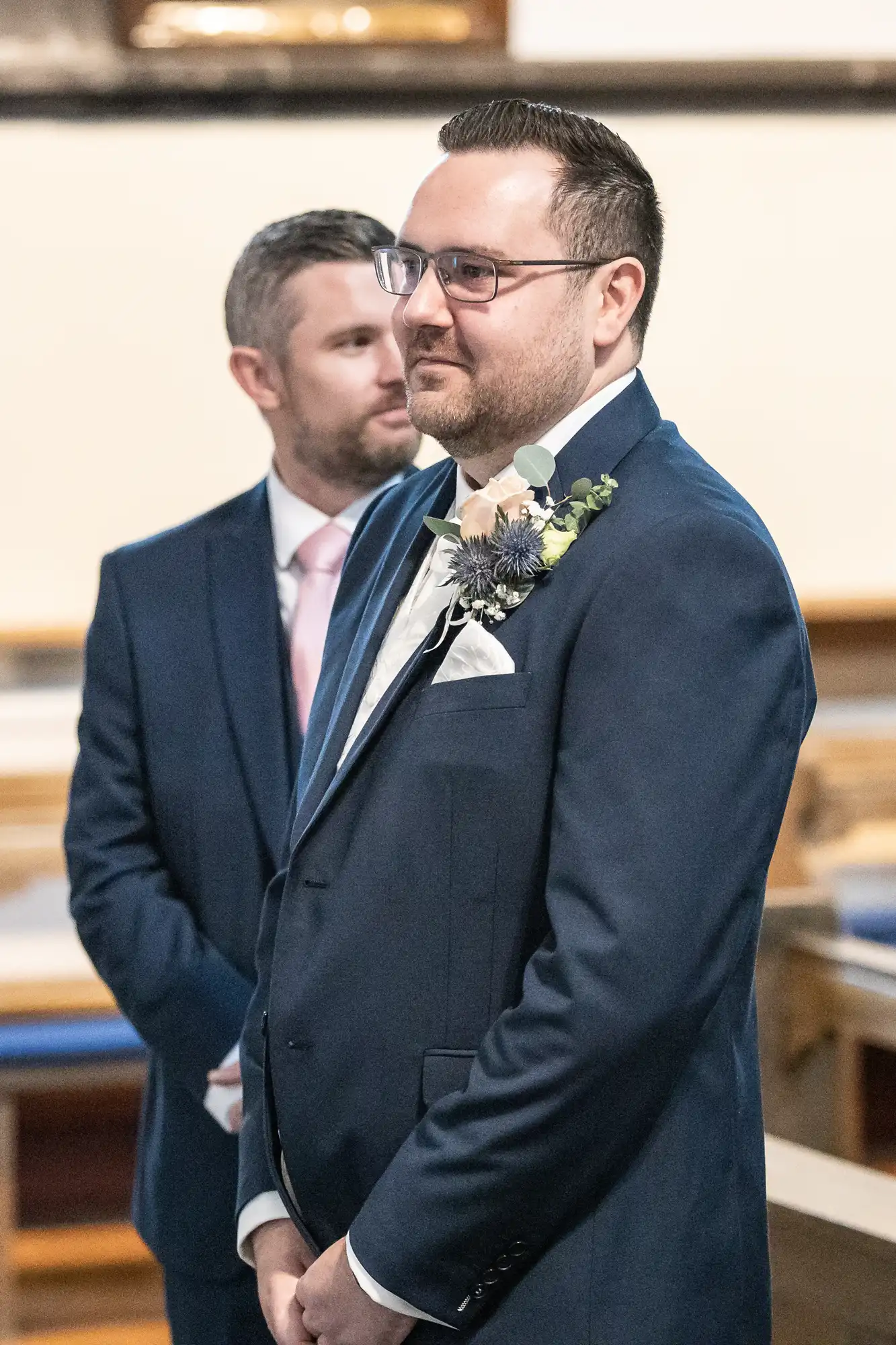 Two men in dark suits stand indoors. The man in front wears glasses, a buttonhole with greenery and cream flowers, and a white pocket square. The man behind him has a short beard and a pink tie. Both look forward, appearing formal and composed.