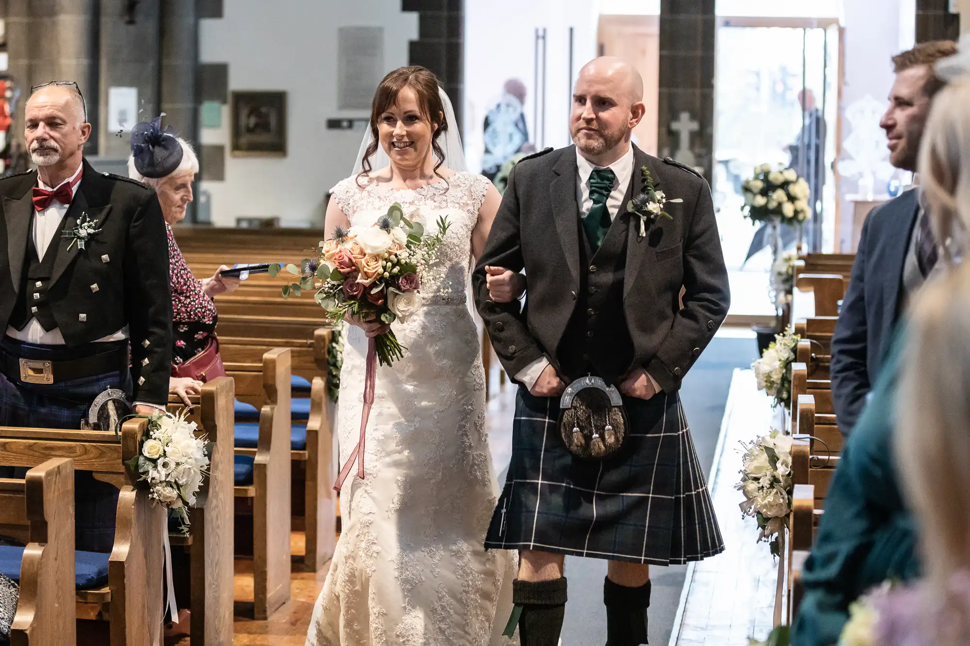 A bride in a white lace dress and veil walks arm-in-arm with a man in a traditional Scottish kilt and jacket down a church aisle lined with flowers, while guests seated and standing look on and smile.