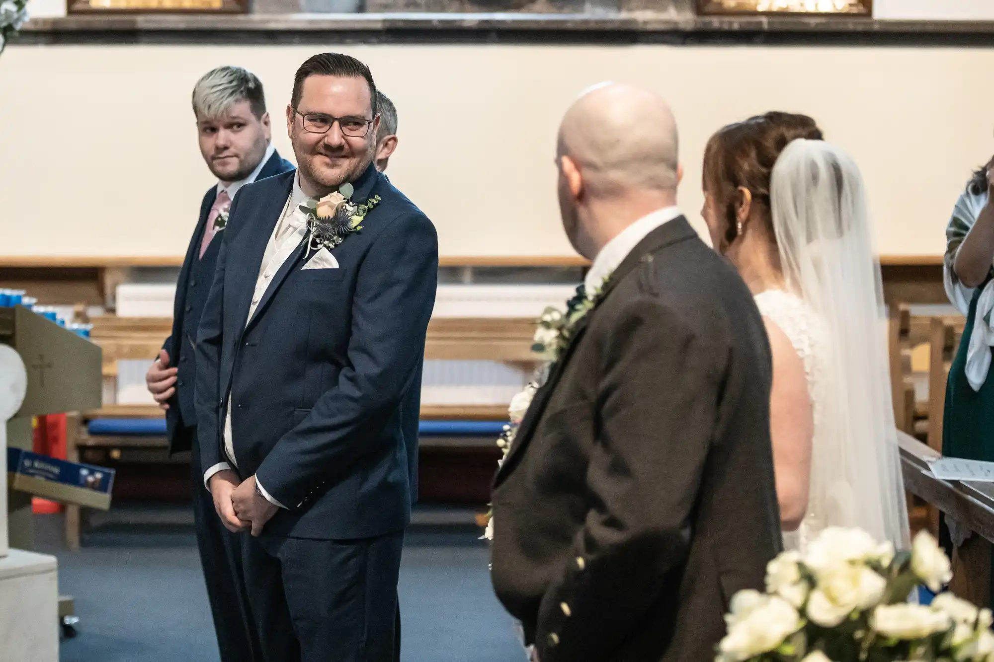 A groom in a blue suit smiles at the bride, who stands beside a man in a dark suit. The bride wears a white dress and veil. Another man with blond hair stands by the groom. The setting is a church decorated with flowers.