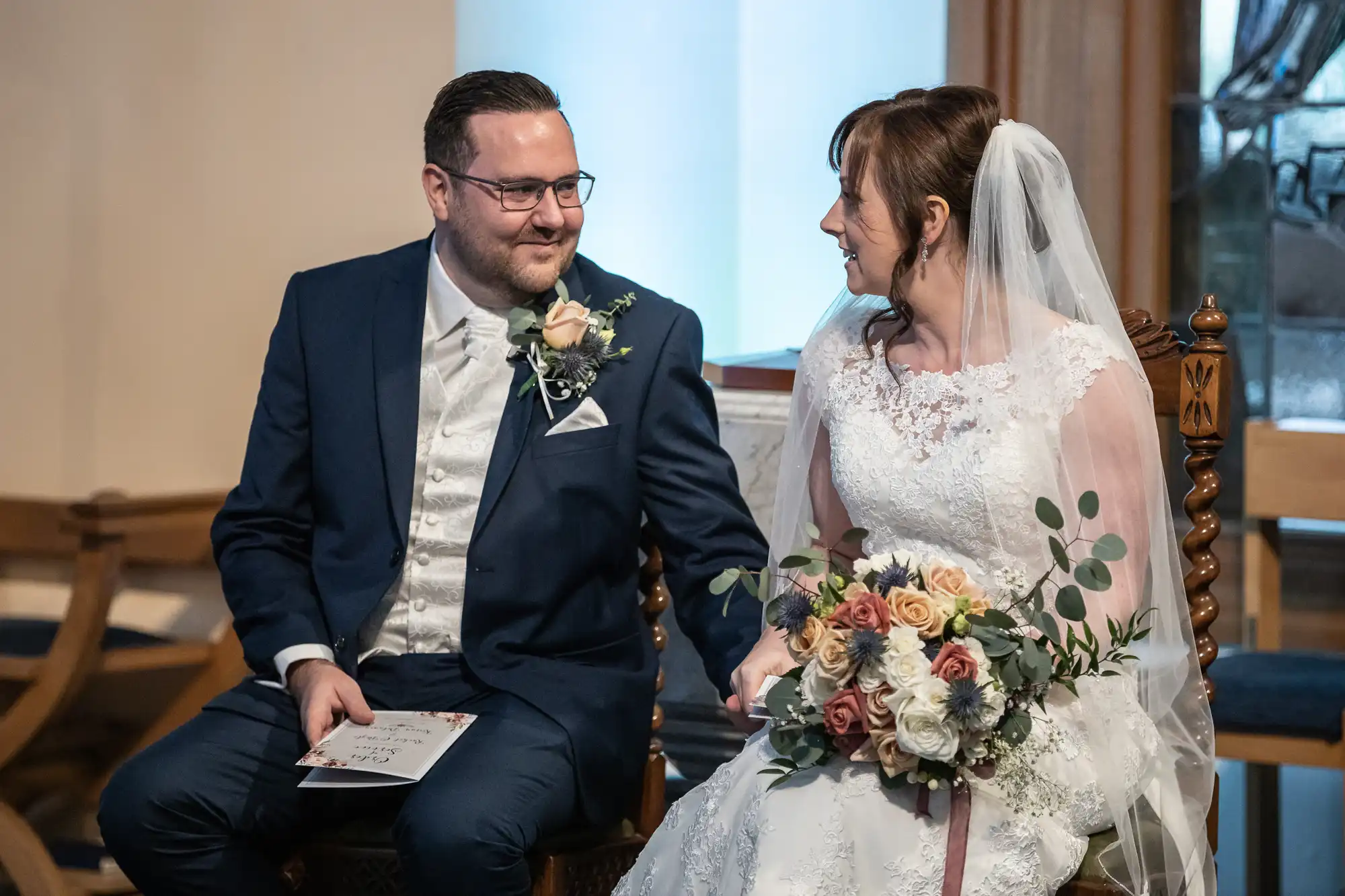 A bride in a white lace gown and veil holds a bouquet of roses and greenery, smiling at the groom beside her in a navy suit with a buttonhole. They sit indoors, holding hands and looking at each other lovingly during their wedding ceremony.