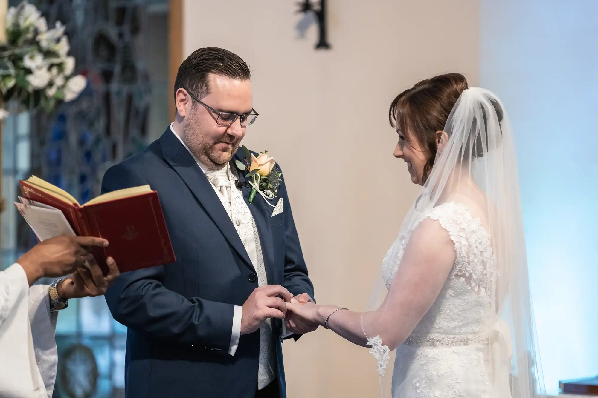 A groom in a navy suit places a ring on the bride’s finger during a wedding ceremony. The bride, wearing a white lace dress and veil, smiles at him. An officiant holds an open red book. They stand indoors with flowers and stained glass in the background.
