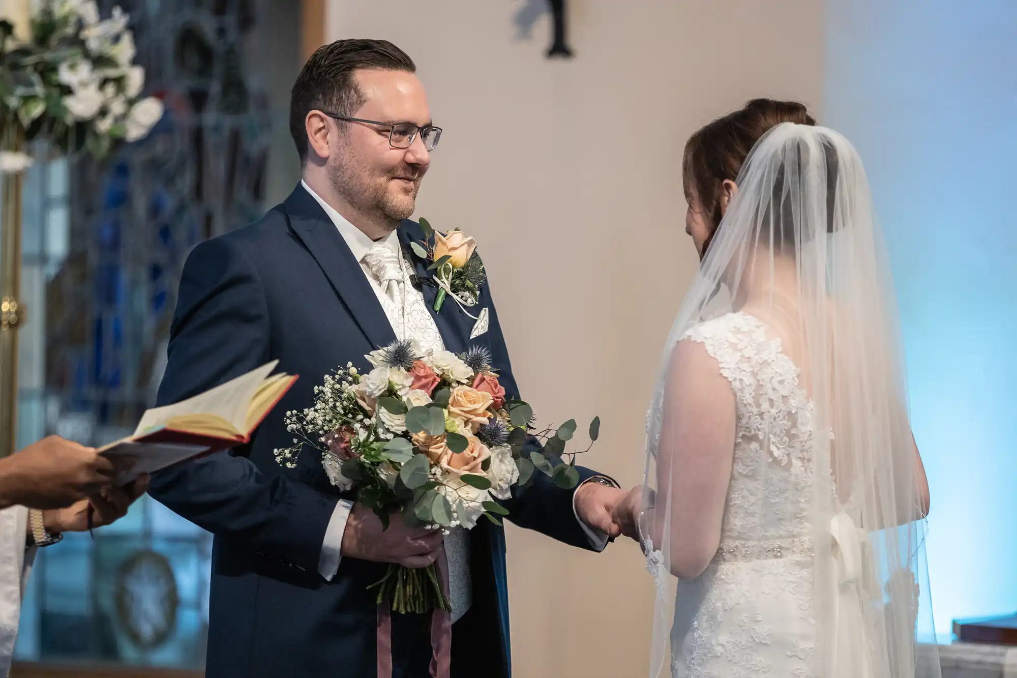 A groom in a navy suit holds a bouquet and smiles at a bride in a white lace gown and veil. They stand facing each other, holding hands during a wedding ceremony. An officiant with a book is partially visible on the left. Background shows soft lighting and stained glass.