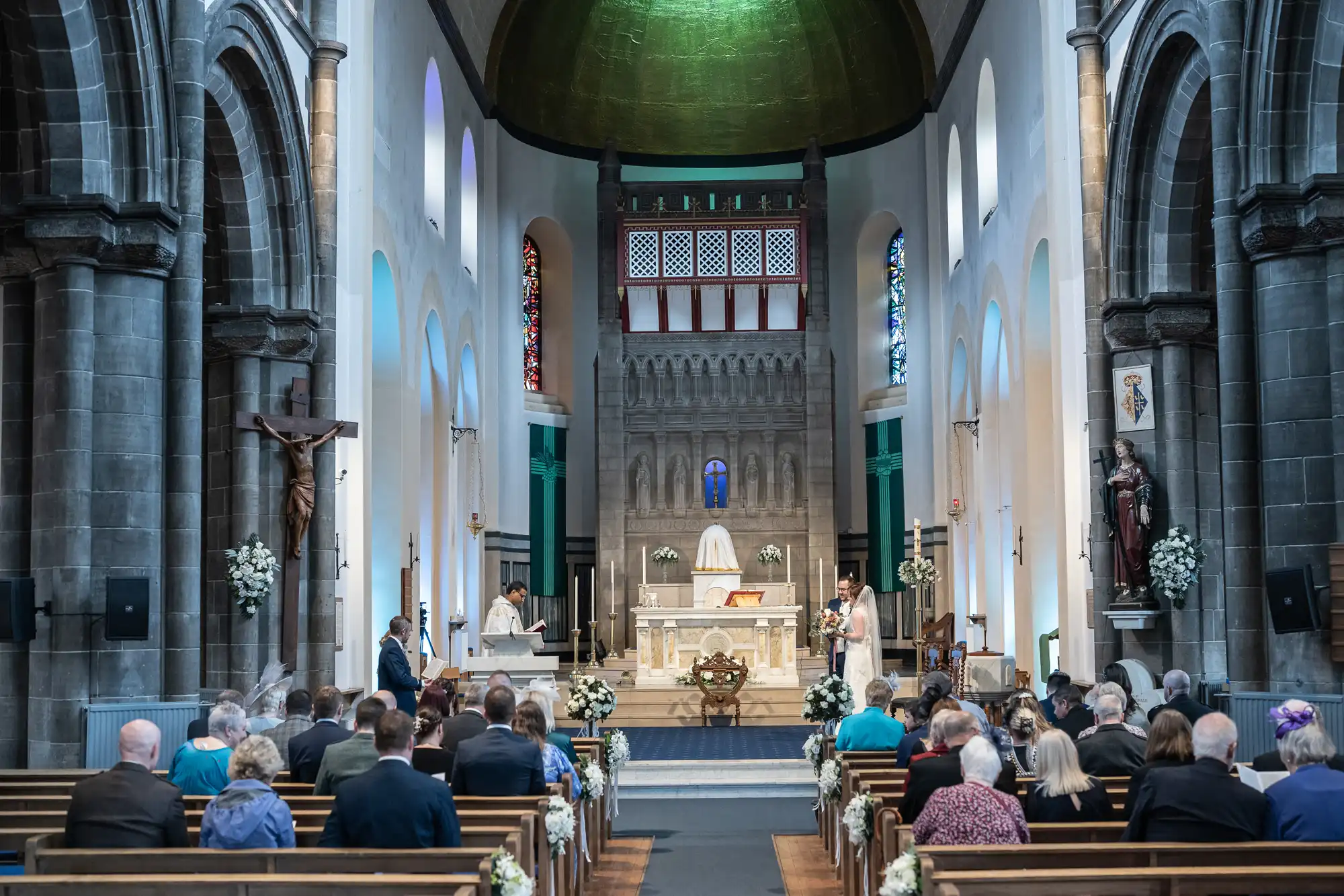 A congregation sits in pews facing an ornate church altar during a wedding ceremony. The church interior features high arches, stained glass windows, statues, and floral decorations. The bride and groom stand at the altar with officiants nearby.