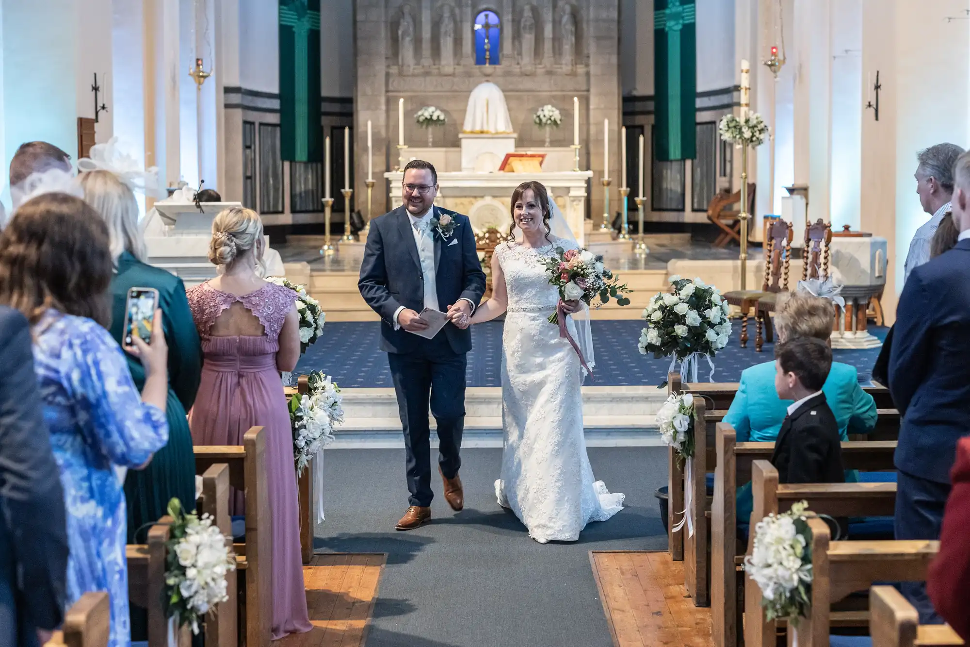 A bride in a white lace dress and a groom in a blue suit walk down the aisle, smiling and holding hands, as wedding guests stand and take photos in a decorated church with floral arrangements and a lit altar in the background.