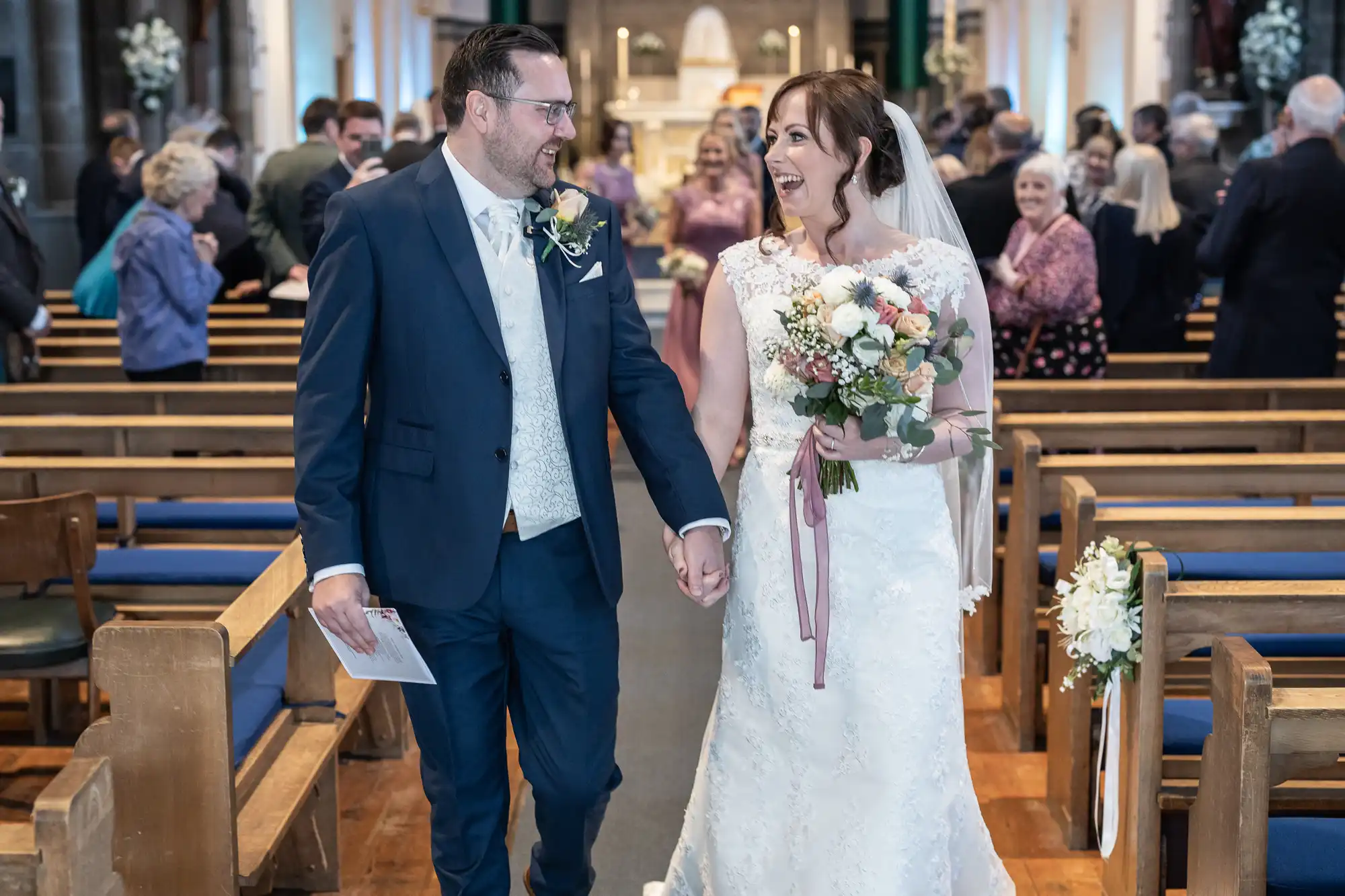 A bride and groom smile joyfully as they walk hand-in-hand down the aisle in a church, surrounded by guests. The groom wears a dark blue suit, while the bride is in a white lace gown holding a bouquet of flowers. People watch and take photos in the background.