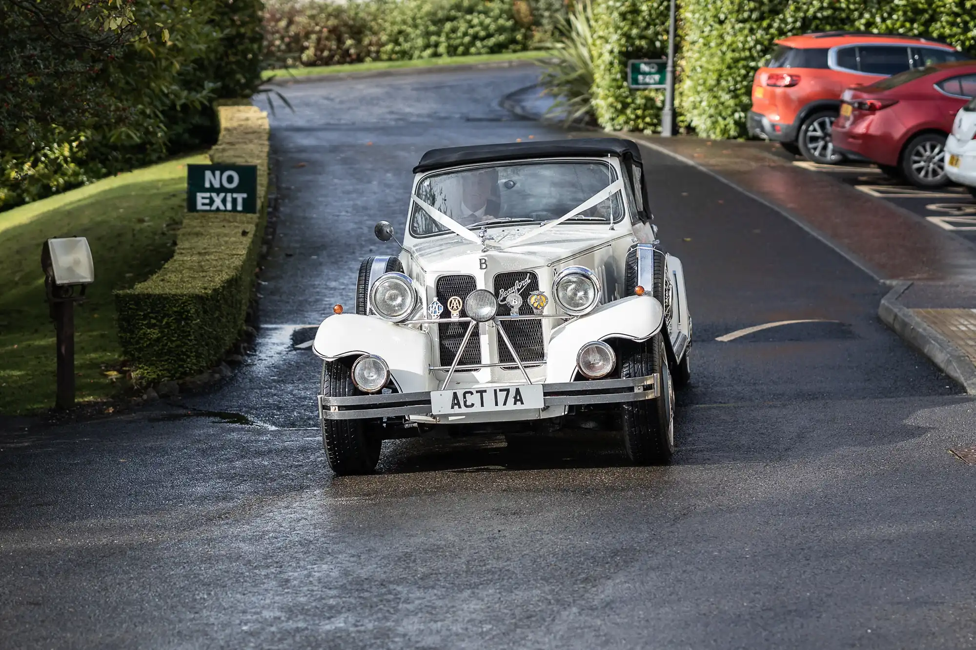 A vintage white wedding car with ribbon decorations is parked on a wet, paved drive. The car has the number plate "ACT 17A." Green hedges, a "NO EXIT" sign, and modern vehicles are visible in the background.