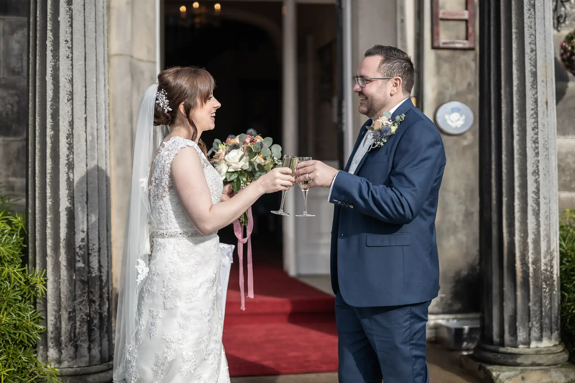 A bride in a white lace dress holding a bouquet and a groom in a blue suit clink champagne glasses and smile at each other outside a building with stone columns and a red carpet leading through an open doorway behind them.