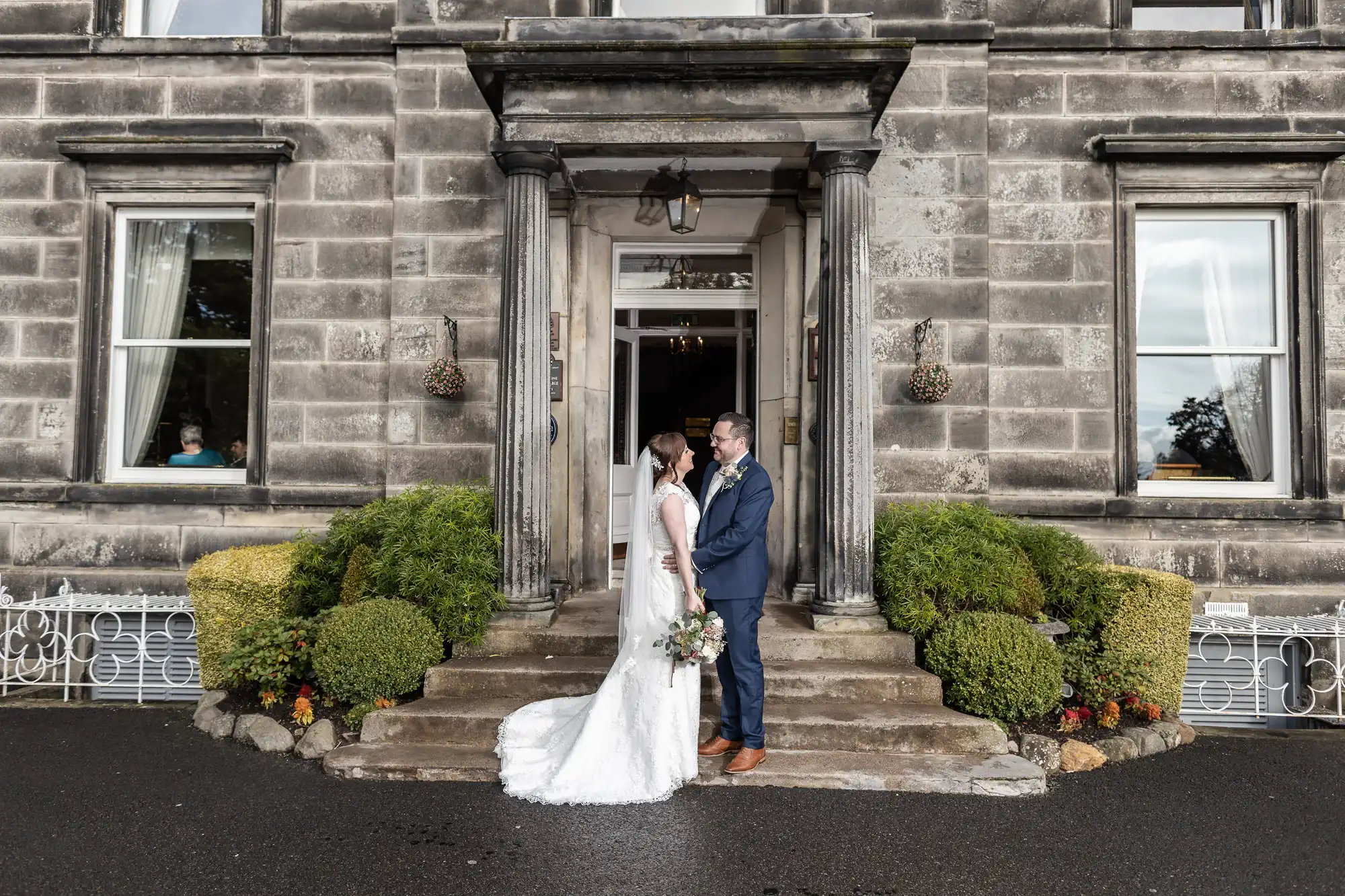 A bride in a white dress and groom in a blue suit stand holding hands on stone steps outside a grand grey stone building with columns, large windows, green bushes, and hanging baskets, gazing at each other on their wedding day.