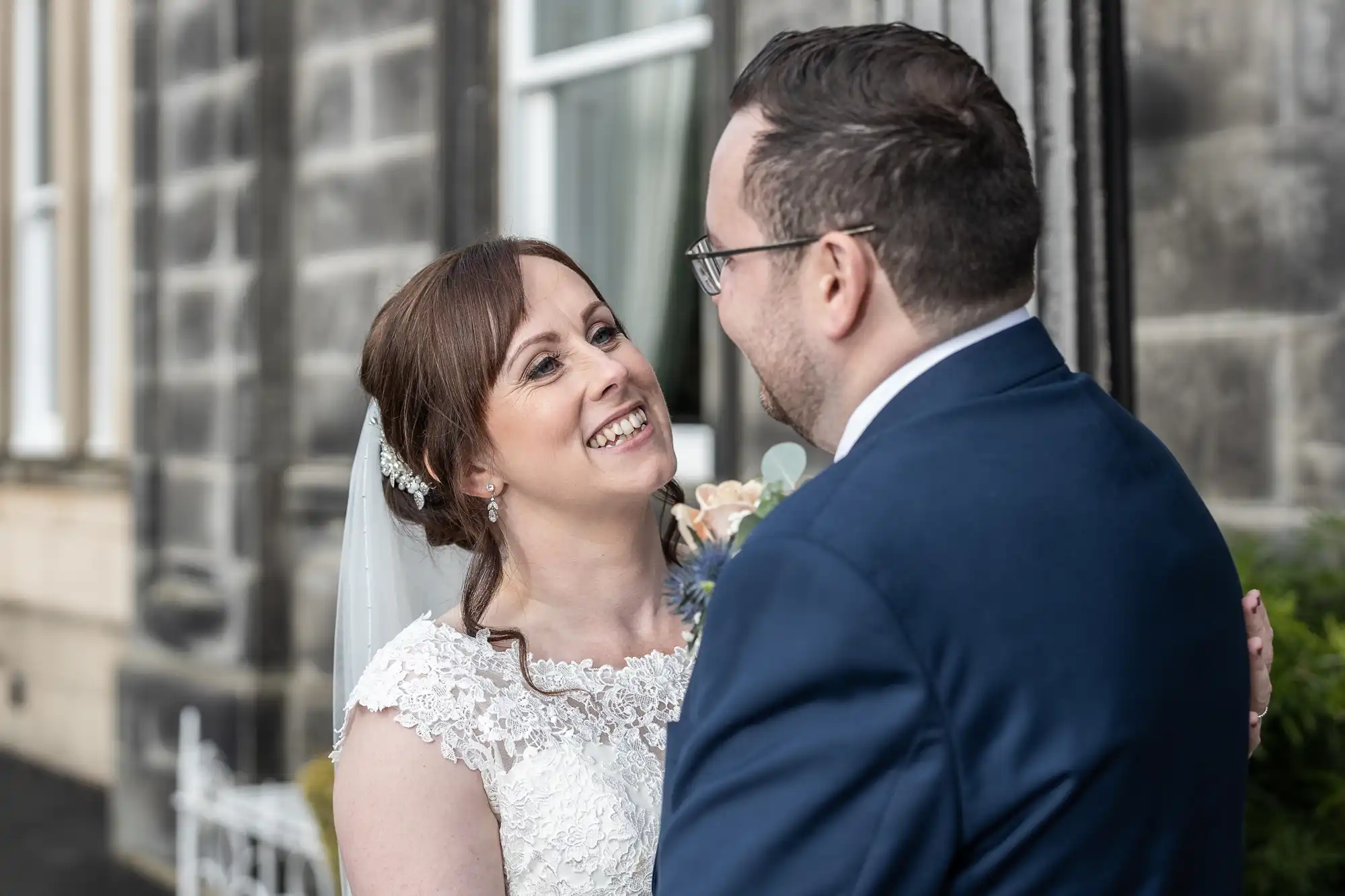 A bride in a lace wedding dress and veil smiles joyfully at her groom, who wears glasses and a navy suit. They stand close together outside a stone building with large windows, sharing a happy, intimate moment on their wedding day.