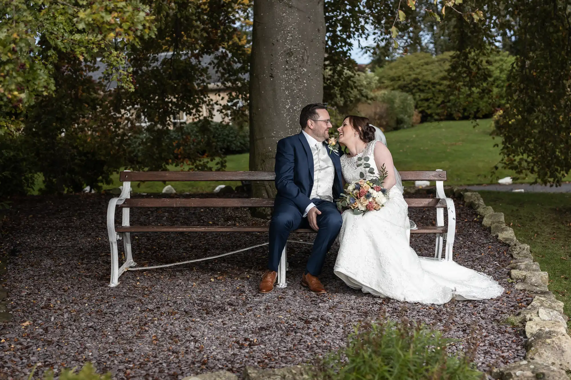 A bride and groom sit closely on a wooden bench outdoors, smiling lovingly at each other. The bride wears a white gown and veil, holding a bouquet, whilst the groom is in a navy suit. They are surrounded by trees and greenery, with a stone border behind them.
