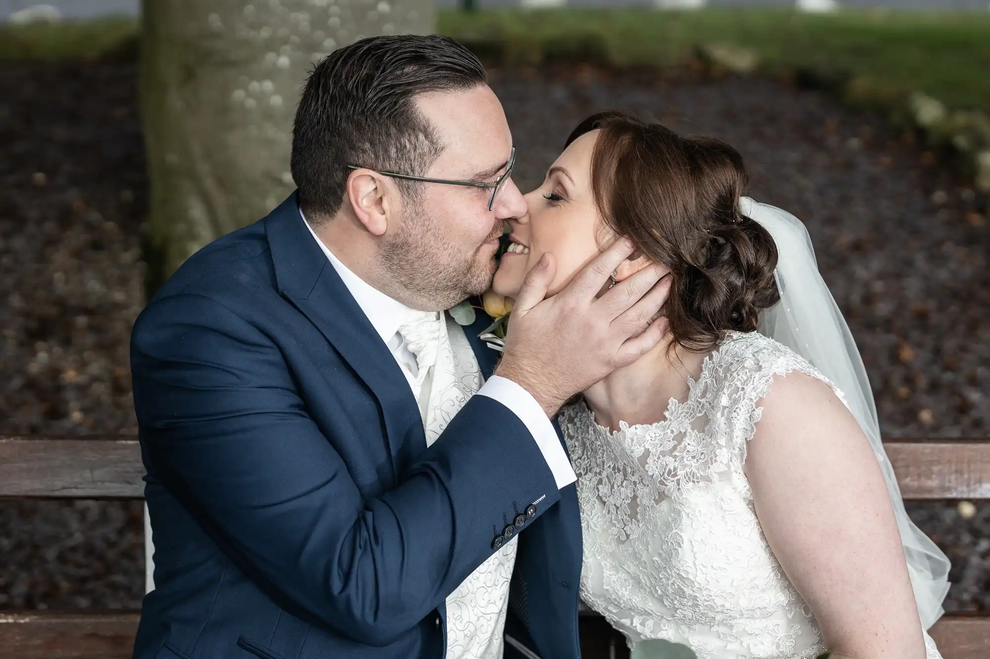 A groom in a navy blue suit and glasses gently kisses a bride in a white lace dress and veil. He holds her chin with one hand as they sit closely on a bench outdoors, sharing a loving, intimate moment on their wedding day.