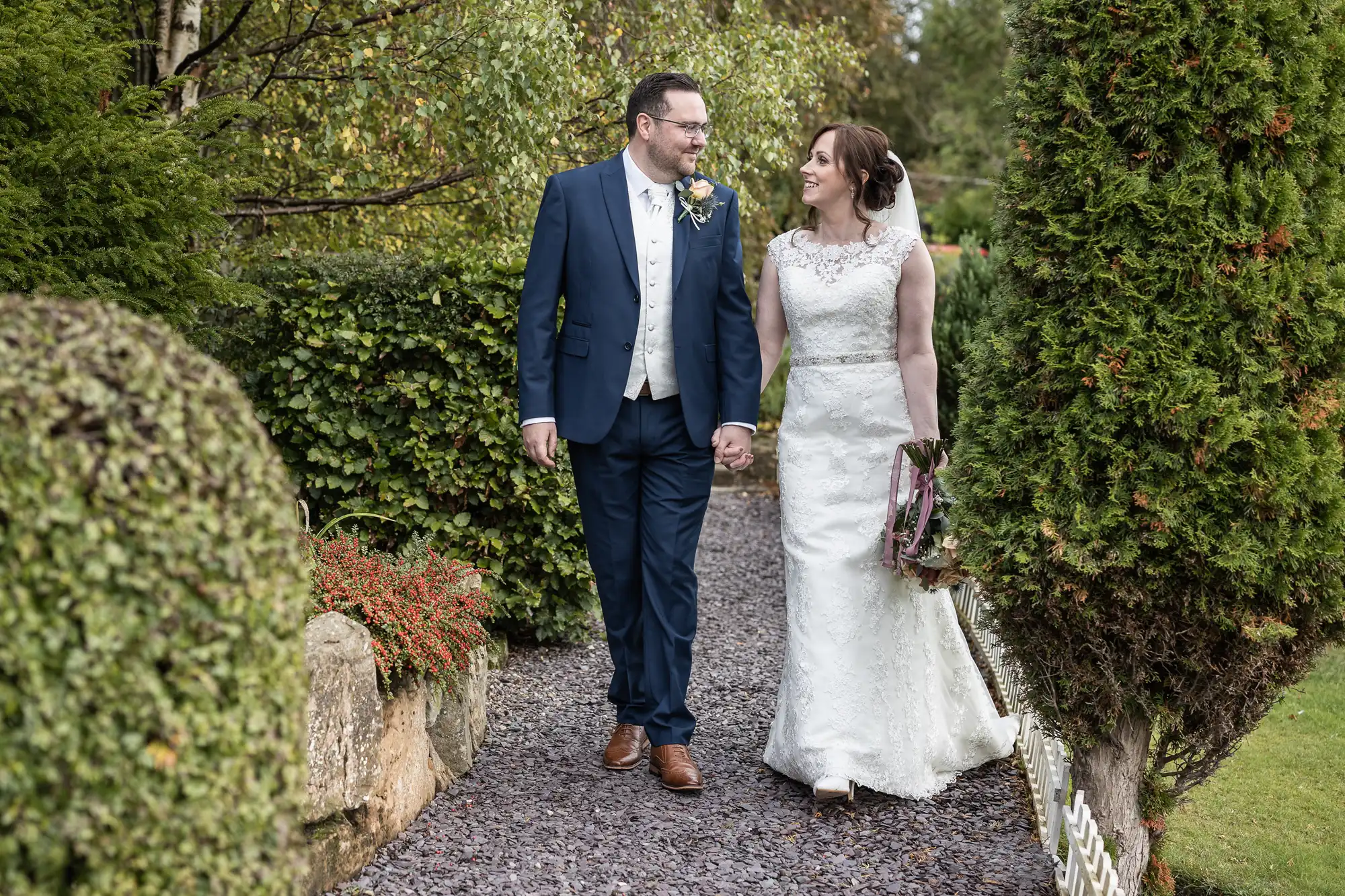 A bride in a white dress and a groom in a blue suit walk hand in hand along a garden path, smiling at each other. Lush greenery and manicured bushes surround them, creating a peaceful and romantic outdoor setting.