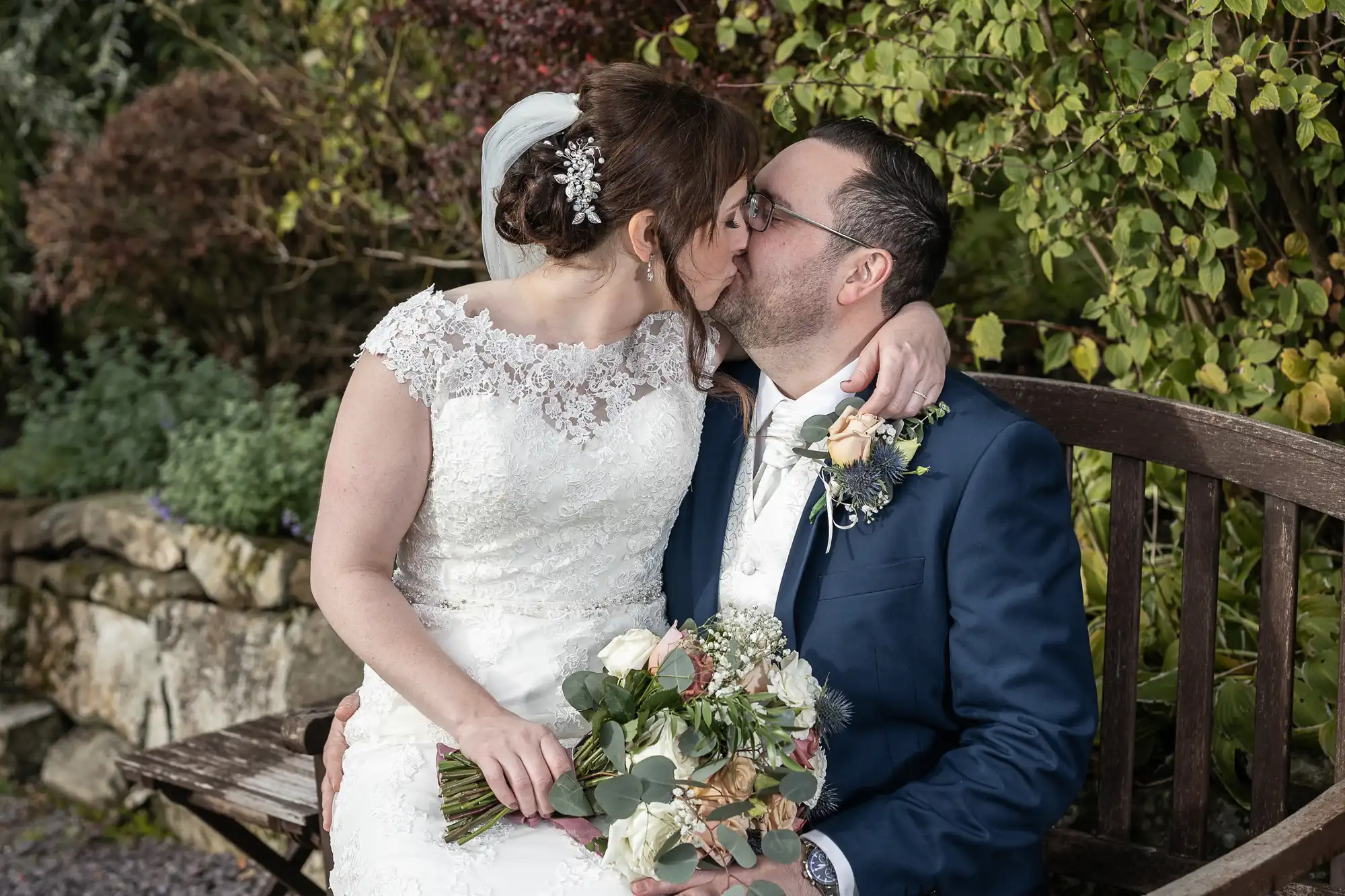 A bride in a white lace wedding dress and a groom in a navy suit sit on a wooden bench, kissing. The bride holds a bouquet of flowers, and greenery and stone walls are visible in the background. Both appear happy and affectionate.