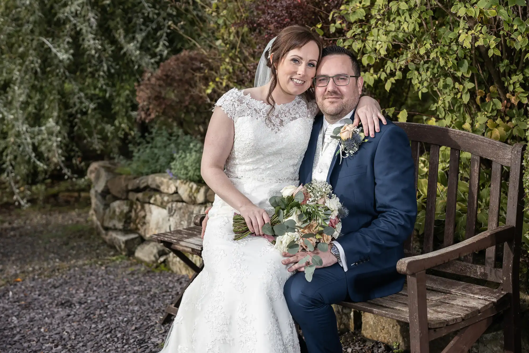 A bride in a white lace dress and veil sits closely beside a groom in a navy suit with a buttonhole, both smiling, on a wooden bench outdoors. The bride holds a bouquet of flowers; greenery and stone wall are in the background.