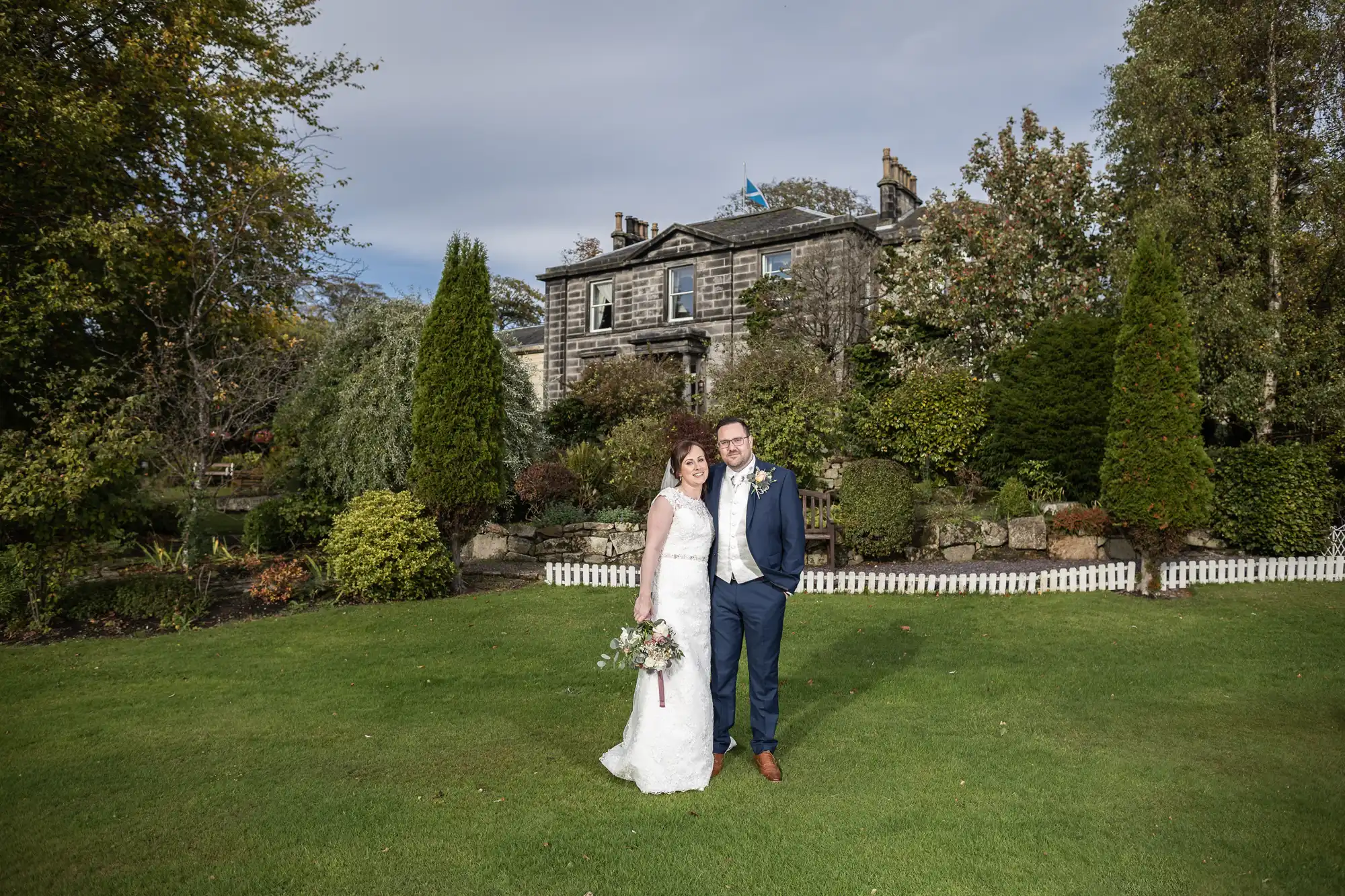 A bride in a white dress holding a bouquet stands beside a groom in a navy suit on a green lawn, smiling. Behind them is a large stone house with white-trimmed windows, surrounded by trees and shrubs on a sunny day.