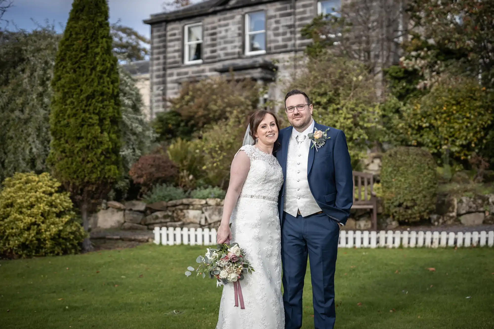A bride in a white lace gown holding a bouquet stands beside a groom in a navy suit and cream waistcoat. They smile in front of a stone house with lush greenery, trees, and a white picket fence in the garden.