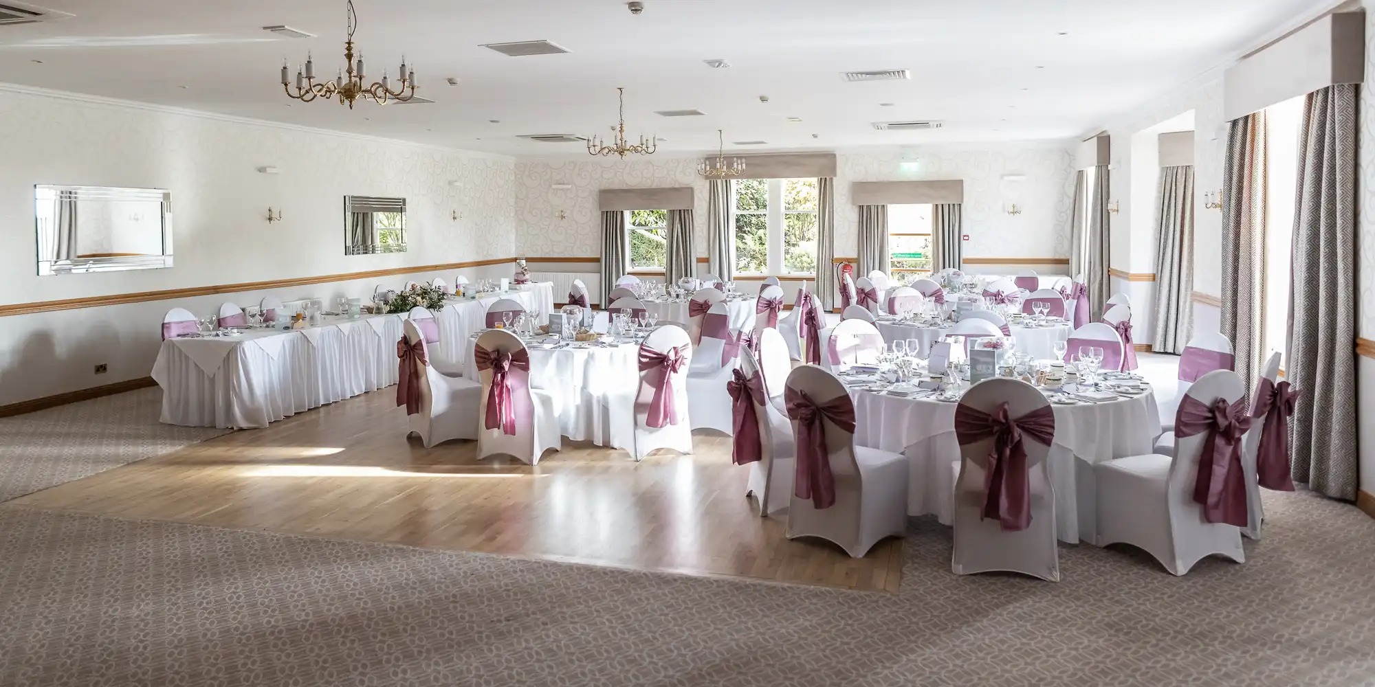 A bright, elegant banquet hall set for an event, with round tables covered in white linen and chairs adorned with mauve bows. Place settings, glassware, and centrepieces decorate the tables. Large windows let in natural light; chandeliers hang from the ceiling.
