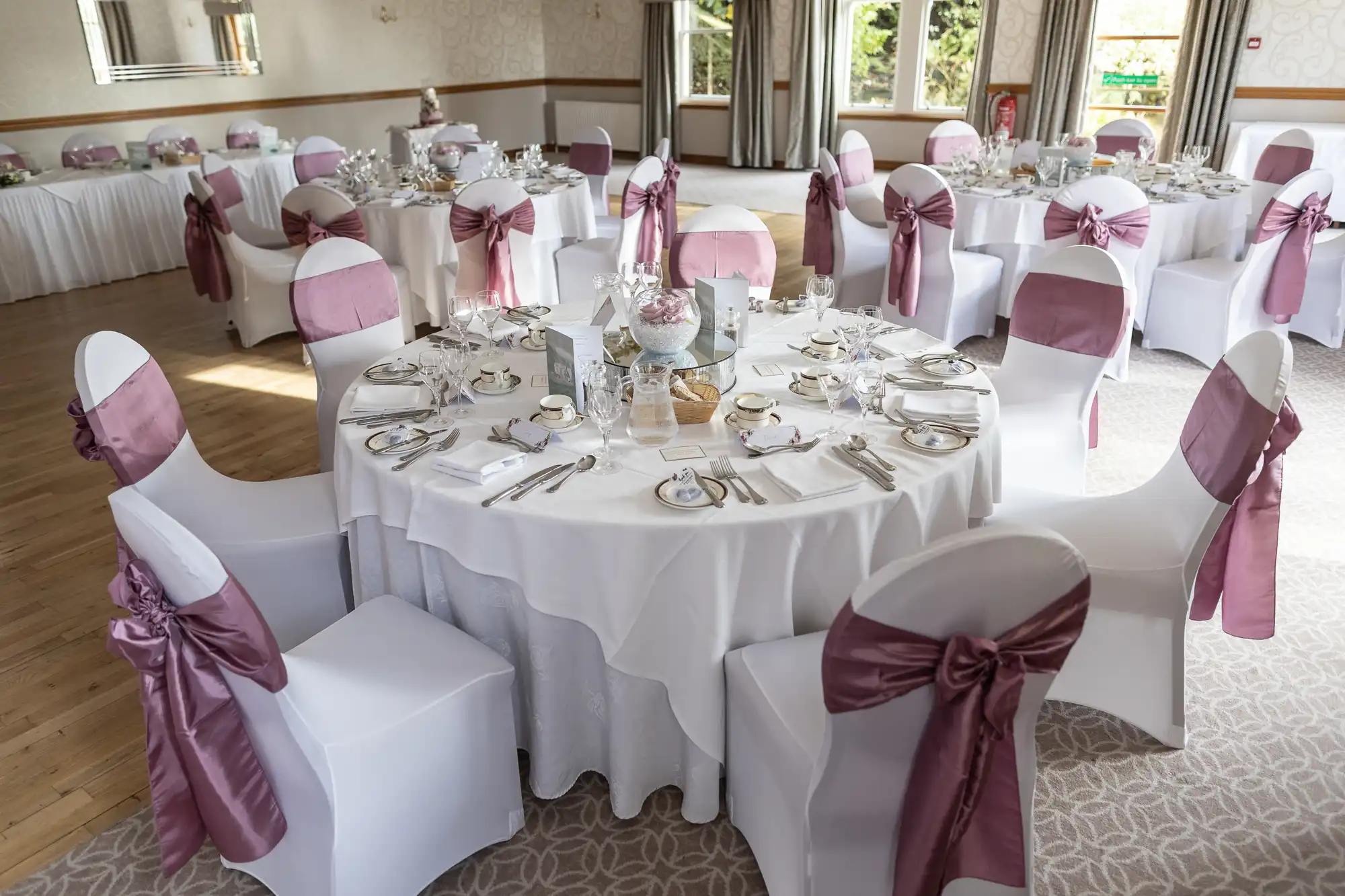 Elegant banqueting hall with round tables set for an event. Tables have white cloths, fine china, glassware, and centrepieces. Chairs are covered in white with mauve sashes. Natural light streams in through large windows with curtains.