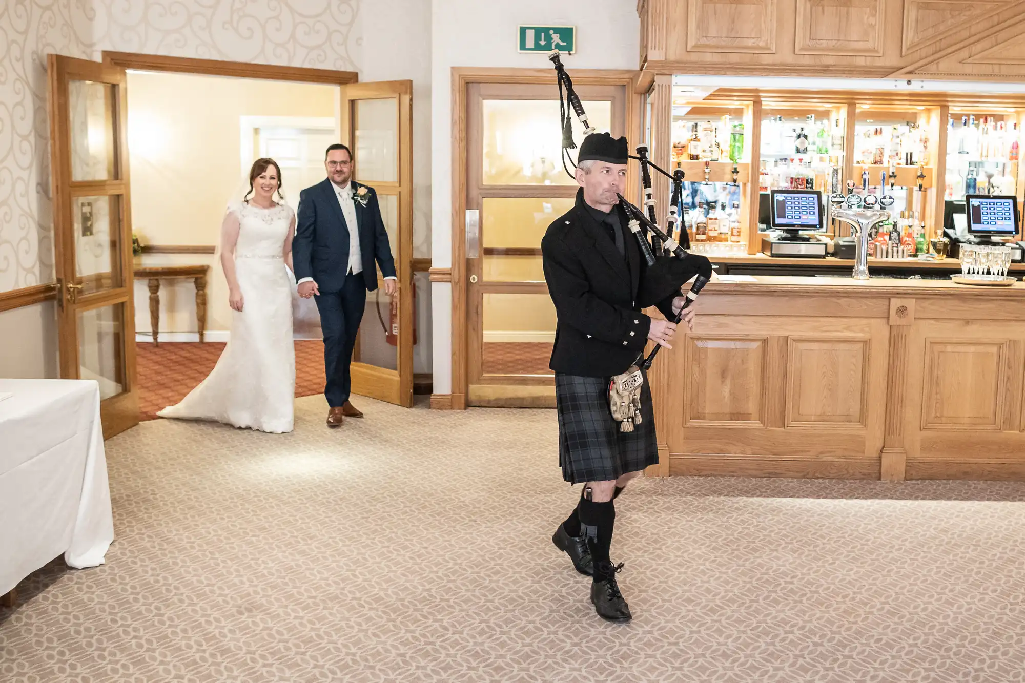 A bride and groom, smiling and holding hands, enter a room as a bagpiper in traditional Scottish attire plays in front of them. The setting appears to be a reception area with a wooden bar and carpeted floor.