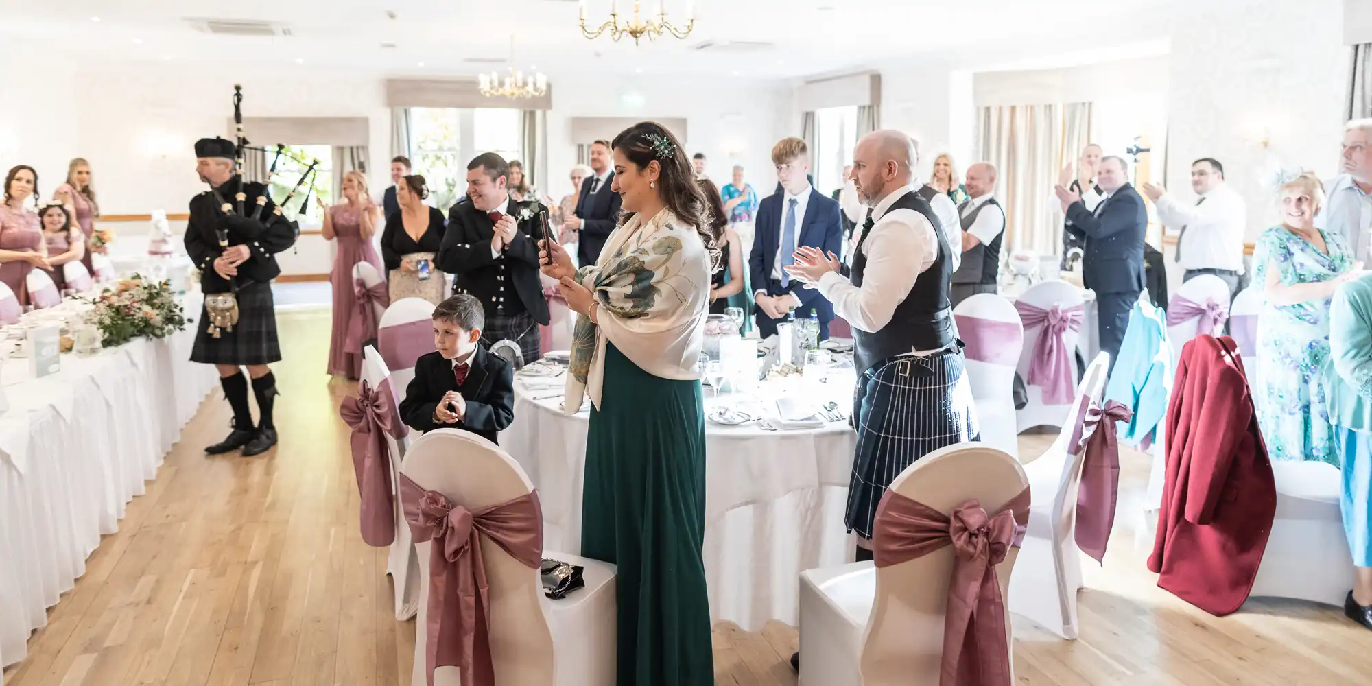 A wedding reception scene with guests standing and applauding around decorated tables. A bagpiper in traditional Scottish attire plays on the left. The room is bright, with white walls and chair covers tied with mauve bows. Everyone is dressed formally.