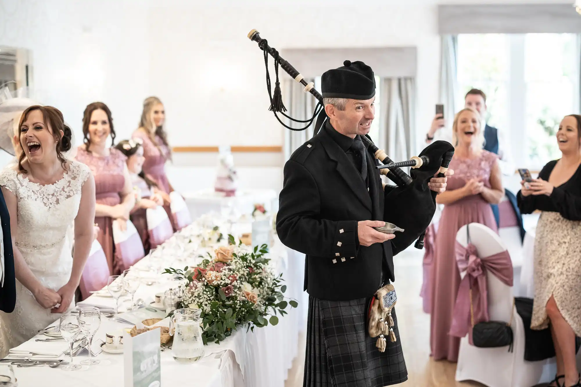 A man in traditional Scottish attire plays bagpipes at a joyful indoor event. Bridesmaids in pink dresses and other guests, some laughing and clapping, flank a long, decorated table with white linen and green floral arrangements. The atmosphere is festive and cheerful.