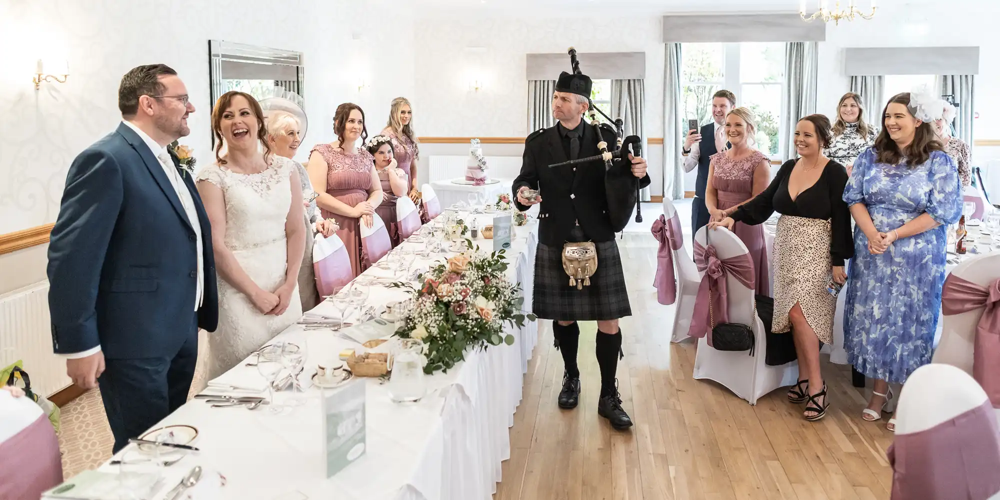 A bride and groom smile at each other beside a long table decorated with flowers. A bagpiper in traditional Scottish dress plays while guests, dressed in semi-formal attire, stand and watch, smiling in a bright, elegant wedding reception hall.