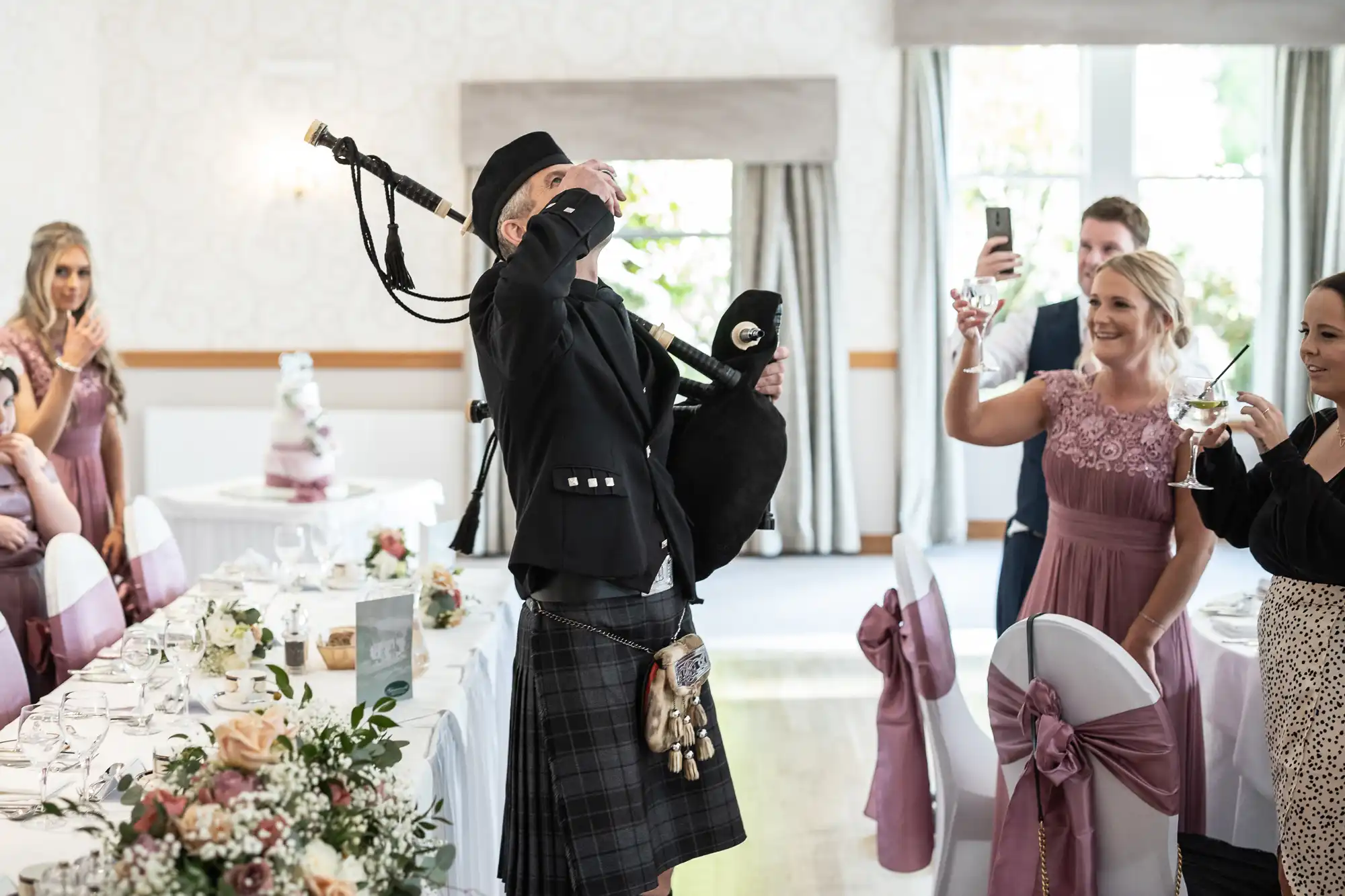 A bagpiper in traditional Scottish attire drinks from a small glass at a wedding reception. Guests in formal dresses smile and take photos. Tables are decorated with flowers, and chairs have pink sashes. The room is bright, with large windows and white walls.