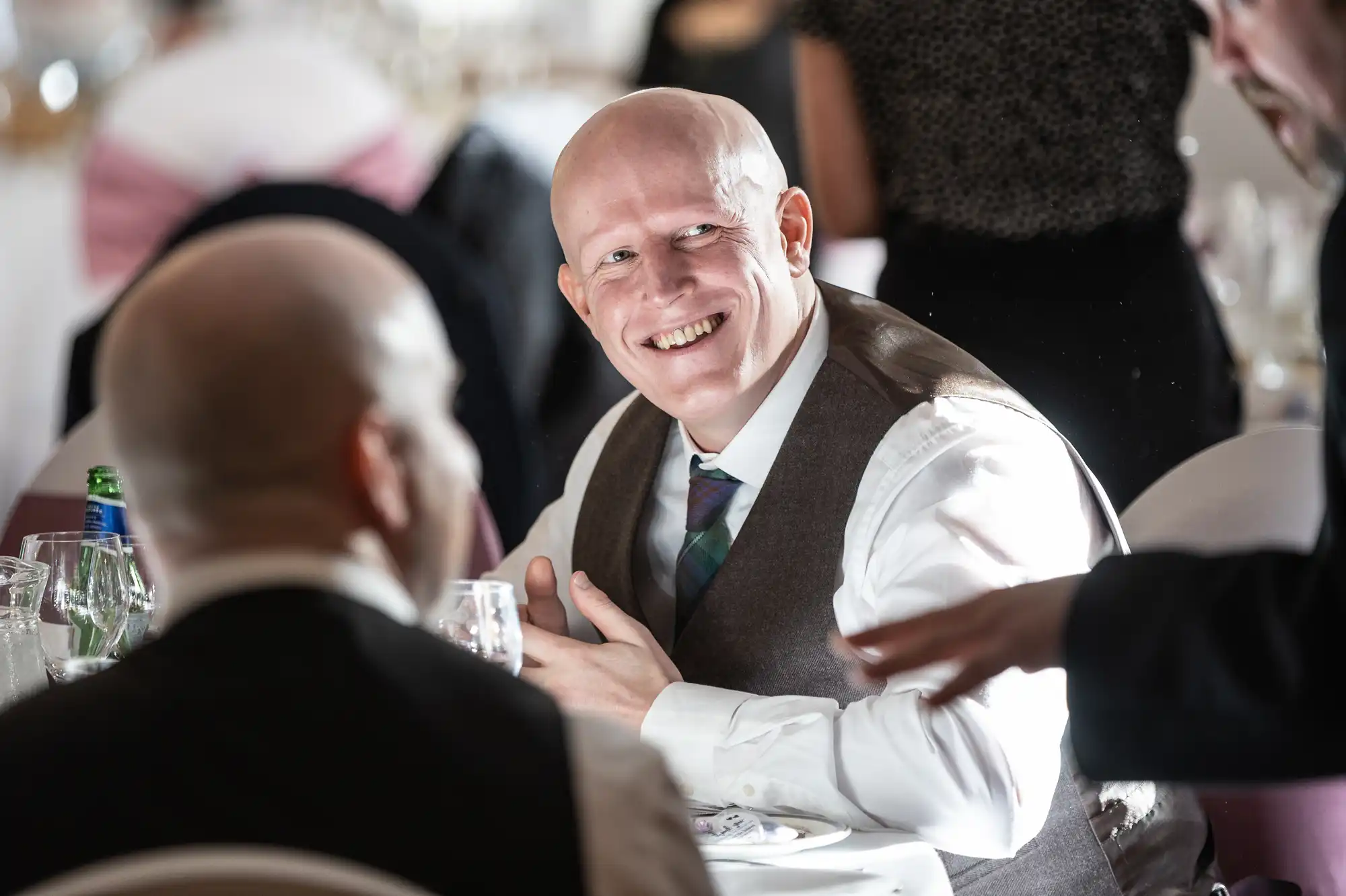 A bald man in a waistcoat and tie smiles warmly whilst sitting at a table set with glasses and plates at a formal event, engaging in lively conversation with other attendees. The atmosphere is bright and festive.
