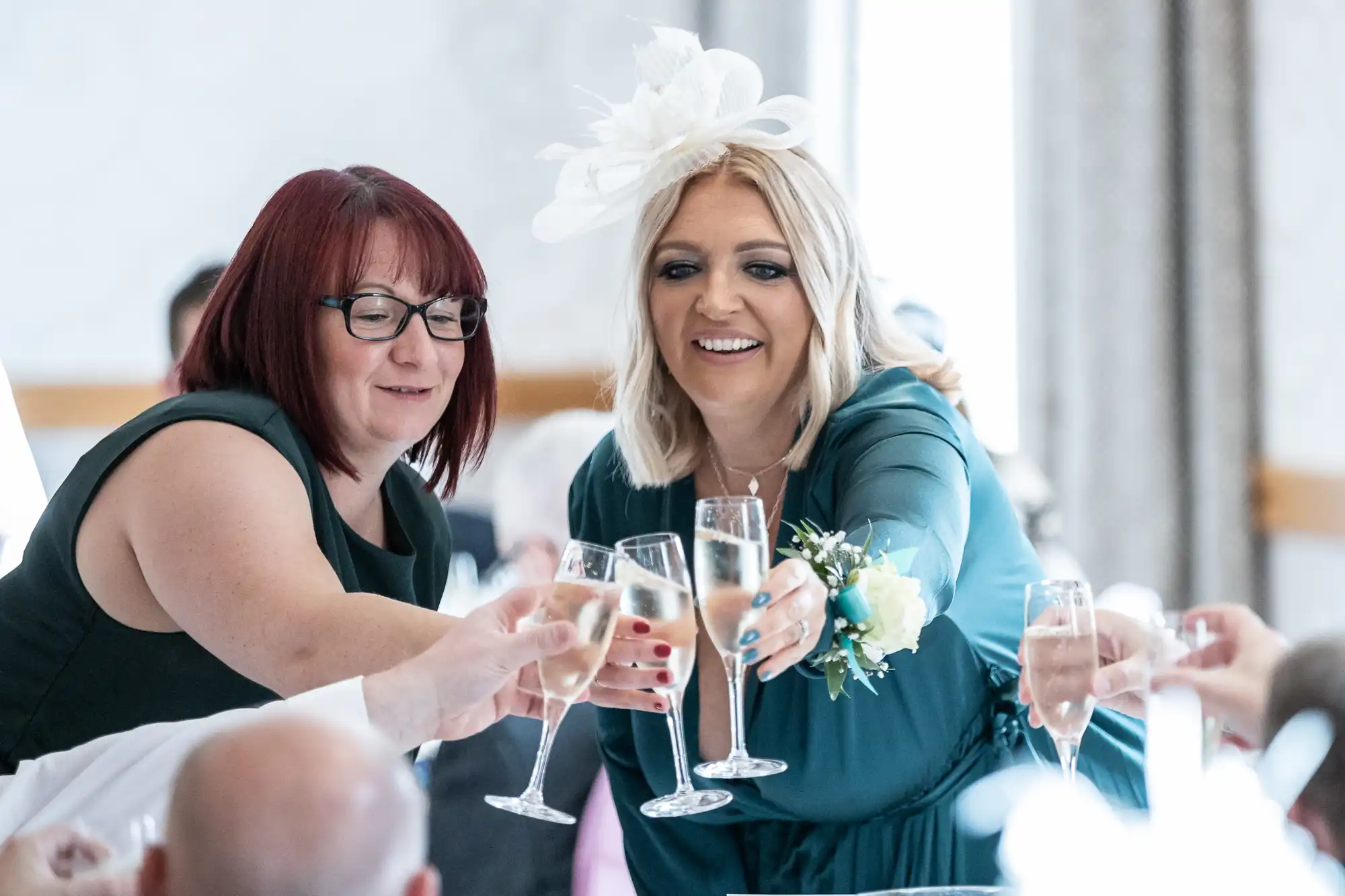 Two women smile and raise champagne glasses for a toast at a formal event. One woman has red hair and glasses, the other has blonde hair and wears a white fascinator headpiece and a green dress with a corsage. Blurred guests are in the foreground.