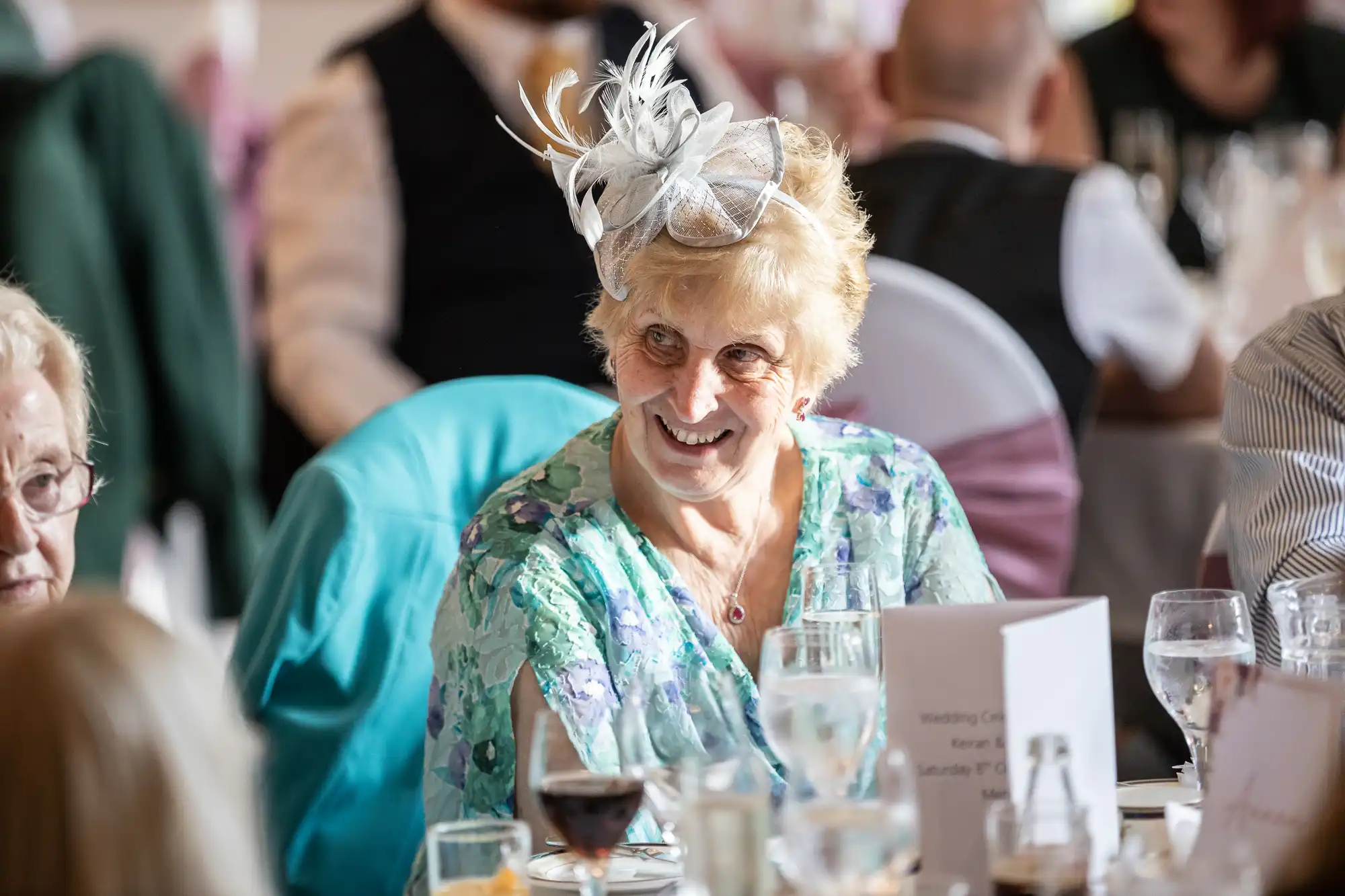 An older woman wearing a light blue floral dress and a decorative white fascinator smiles whilst sitting at a table set with glasses and menus at a formal event, possibly a wedding. Other guests and blurred tables are visible in the background.