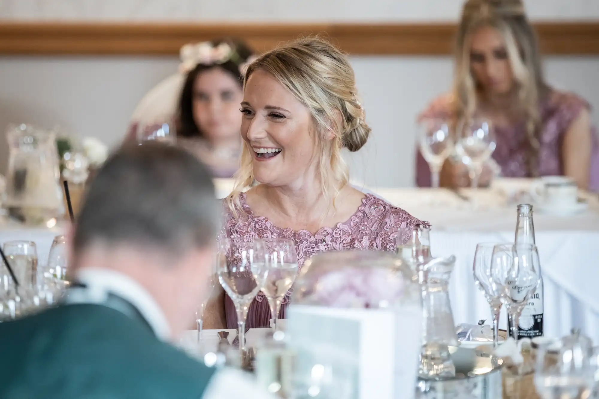 A woman with blonde hair in an updo smiles at a table set for a formal event. She wears a lilac lace dress. Other guests, including women in similar dresses, are visible in the background. Wine glasses, plates, and a fizzy drink bottle are on the table.