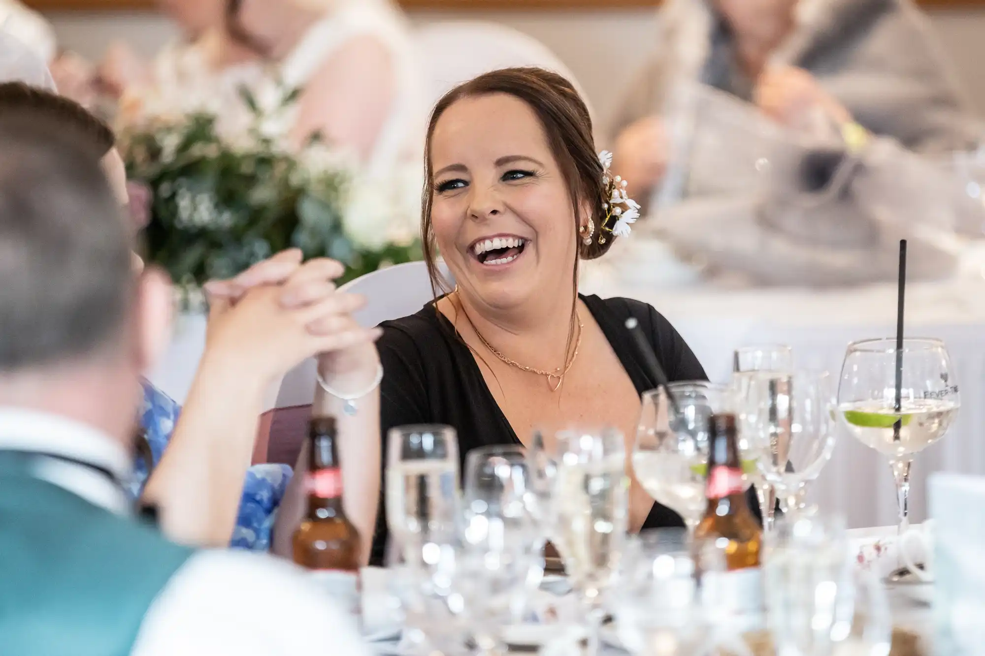 A woman with brown hair in a black dress laughs joyfully at a table set with glasses, bottles, and a lime-garnished drink during a lively social event, surrounded by other guests in a softly lit, elegant indoor setting.