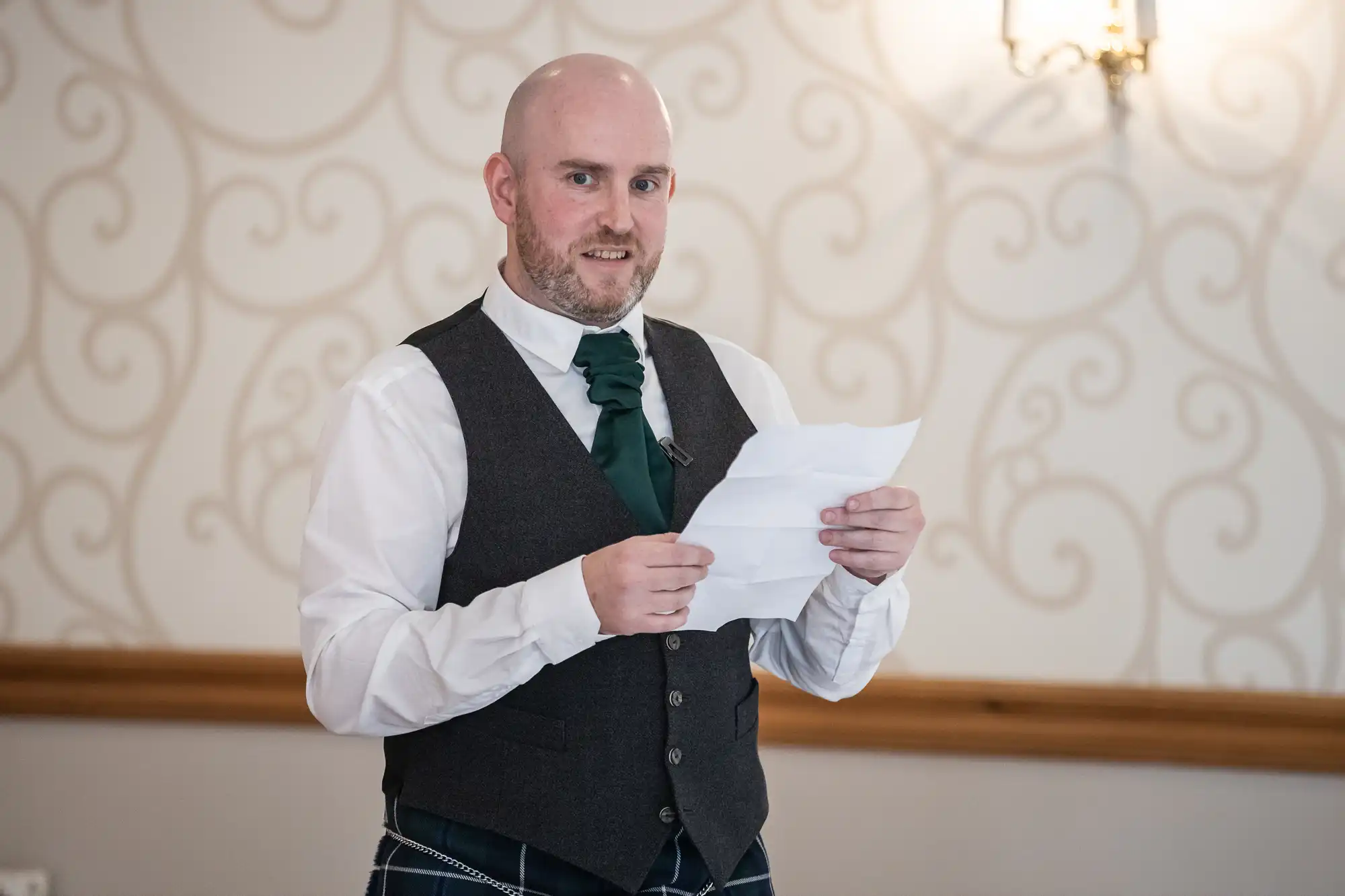 A bald man with a beard, dressed in a white shirt, dark waistcoat, green tie, and tartan kilt, holds and reads from a piece of paper whilst standing indoors against a cream wall with a decorative swirl pattern and a wall lamp.