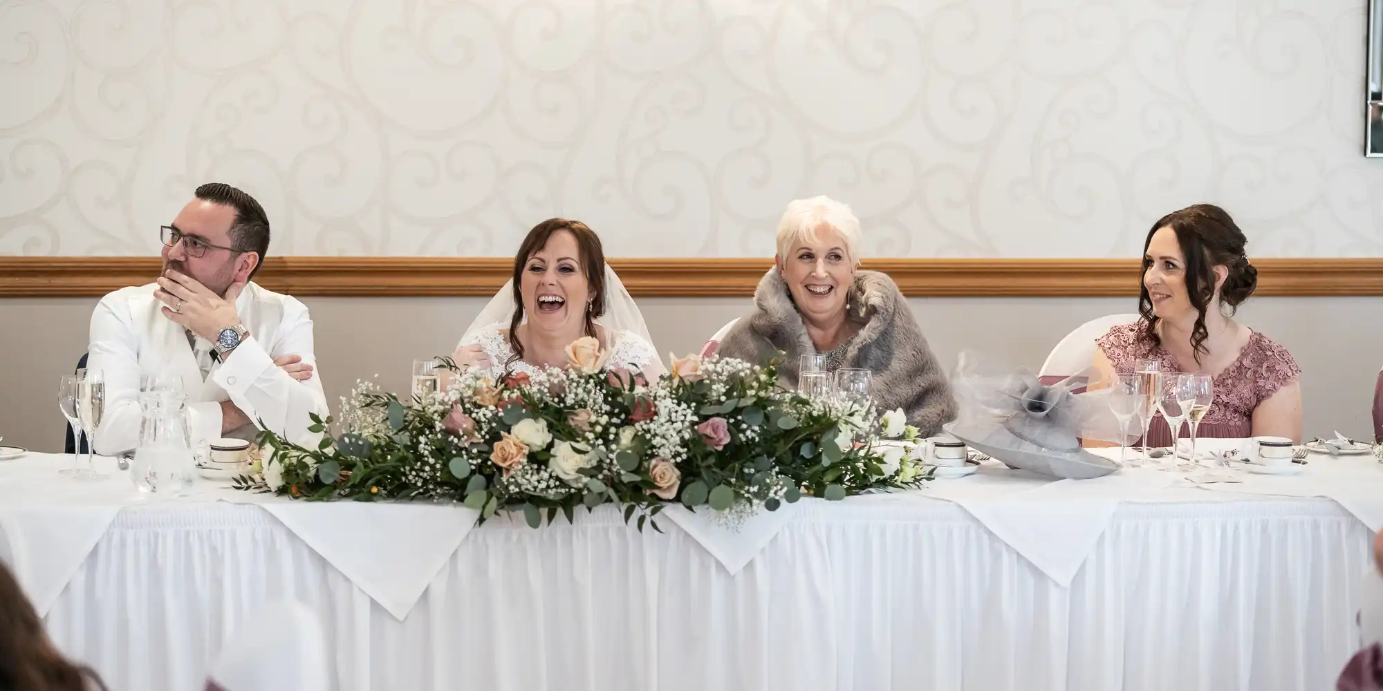 Four people sit at a wedding top table. A bride in white and veil sits beside two women in pink dresses and an older woman in a grey stole. All are smiling and laughing. A floral arrangement decorates the table, which is covered with a white cloth.