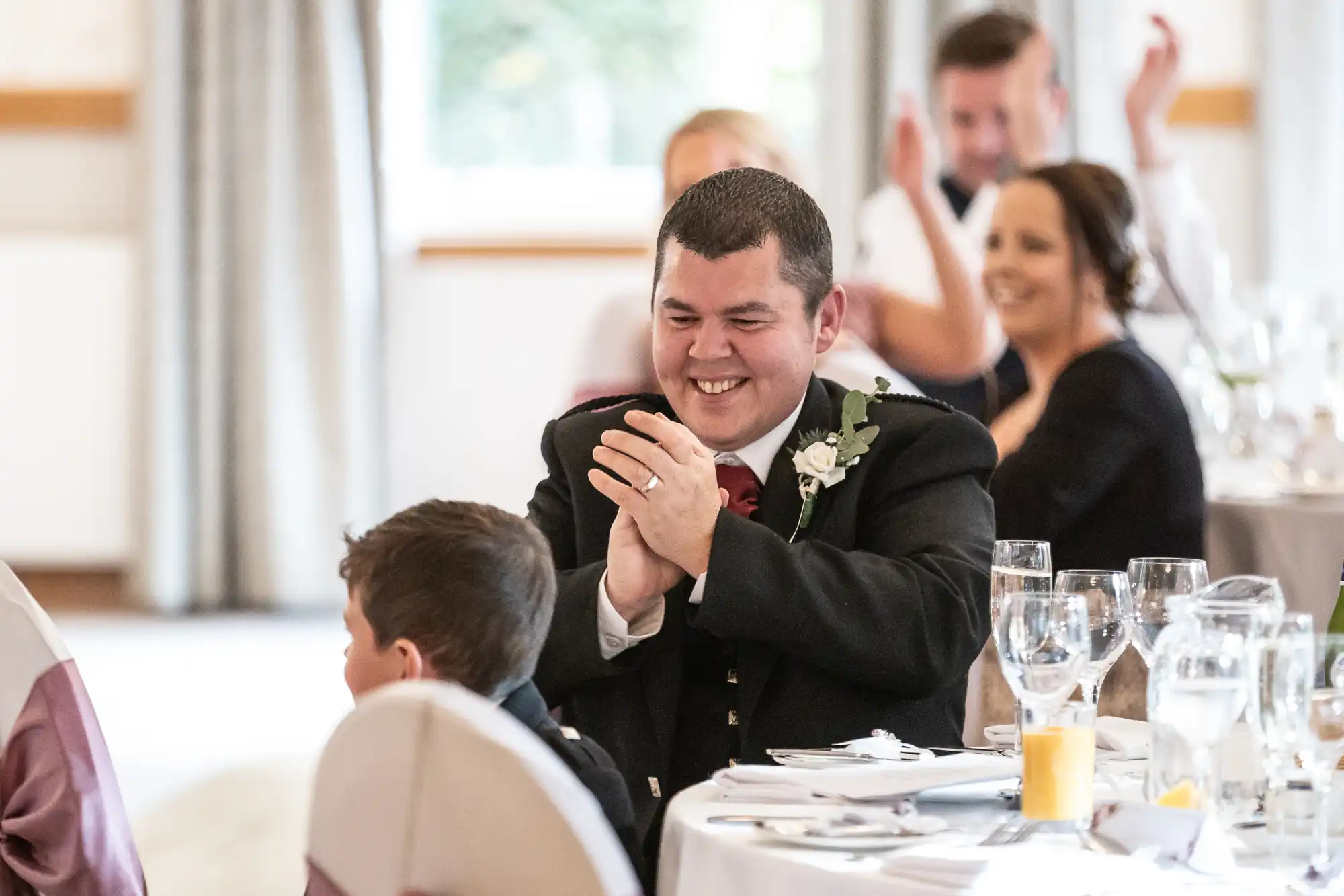A man in formal attire with a buttonhole smiles and claps at a decorated table during a celebration. Several people, including a child, are around him, some also clapping and smiling. Glasses and a juice glass are visible on the table.