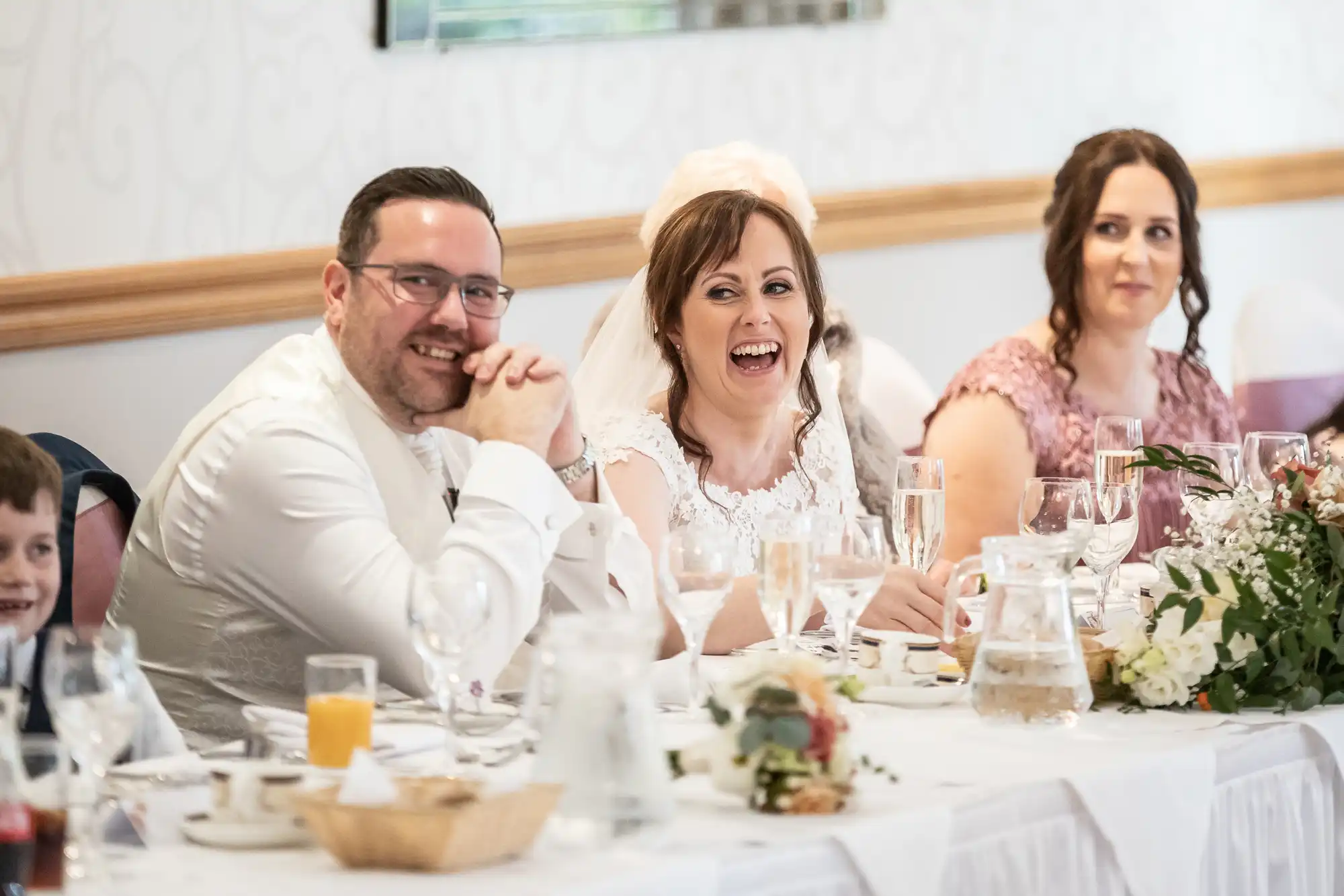 A bride and groom sit smiling at a wedding reception table, joined by guests. The bride is in white with a veil, laughing, whilst the groom in a white shirt and glasses smiles. Floral arrangements, drinks, and place settings are on the white-clothed table.