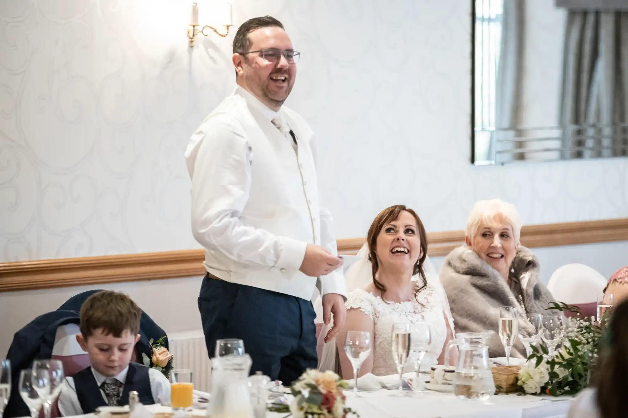 A man in a white waistcoat stands and smiles, giving a speech at a wedding reception table. A woman in a white dress and another woman in a fur stole smile beside him. A young boy with a drink sits at the table, which is decorated with flowers and glassware.