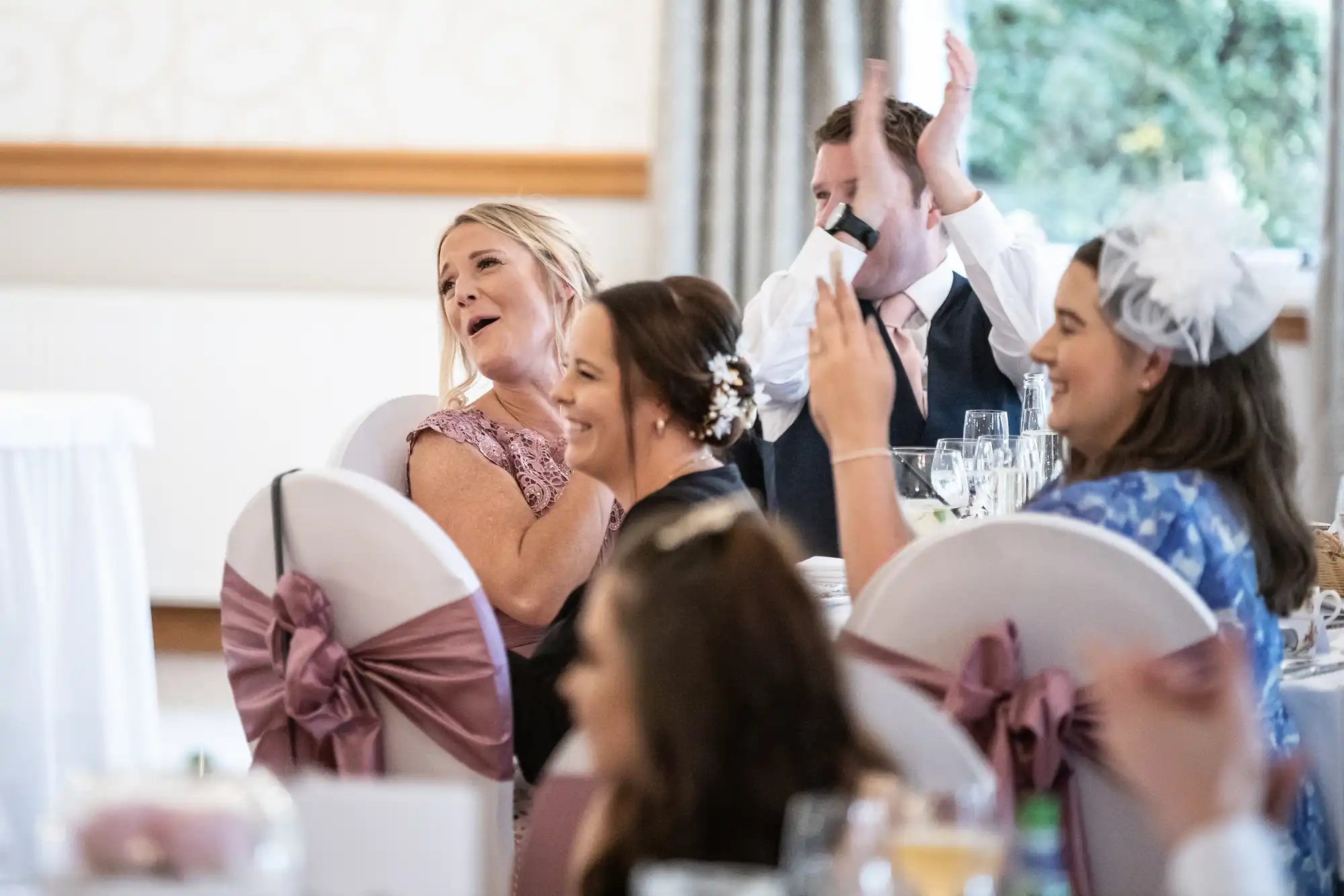 Four guests at a wedding reception sit at a round table decorated with mauve chair sashes. A woman in a mauve dress laughs, another in a hair clip smiles, a man in a waistcoat claps, and a woman in blue with a white fascinator applauds.