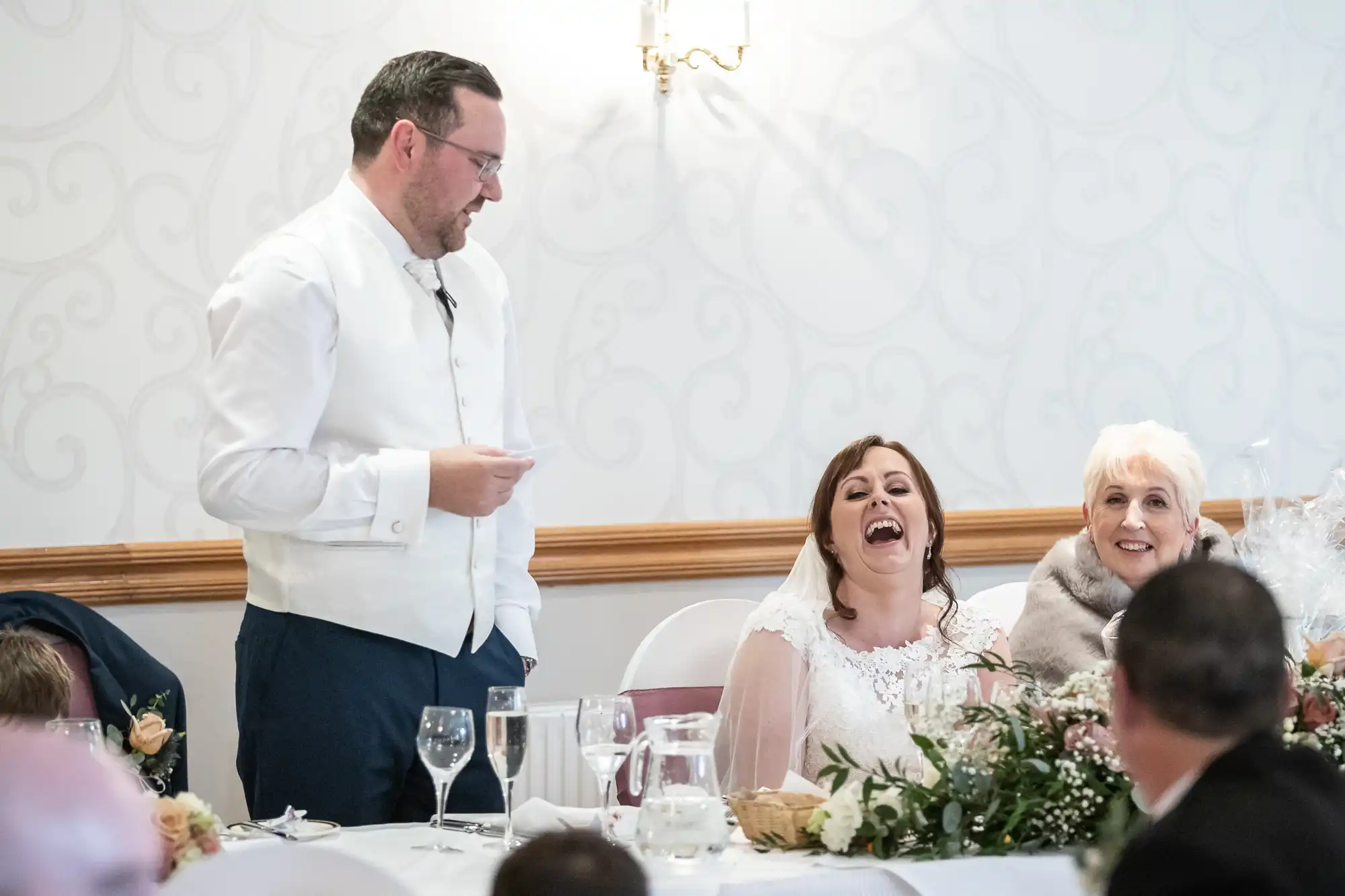 A man in a white waistcoat and glasses stands giving a speech at a wedding table. The bride, wearing a white dress and veil, laughs joyfully. An older woman next to her smiles. The table is set with flowers, glasses, and a water jug.