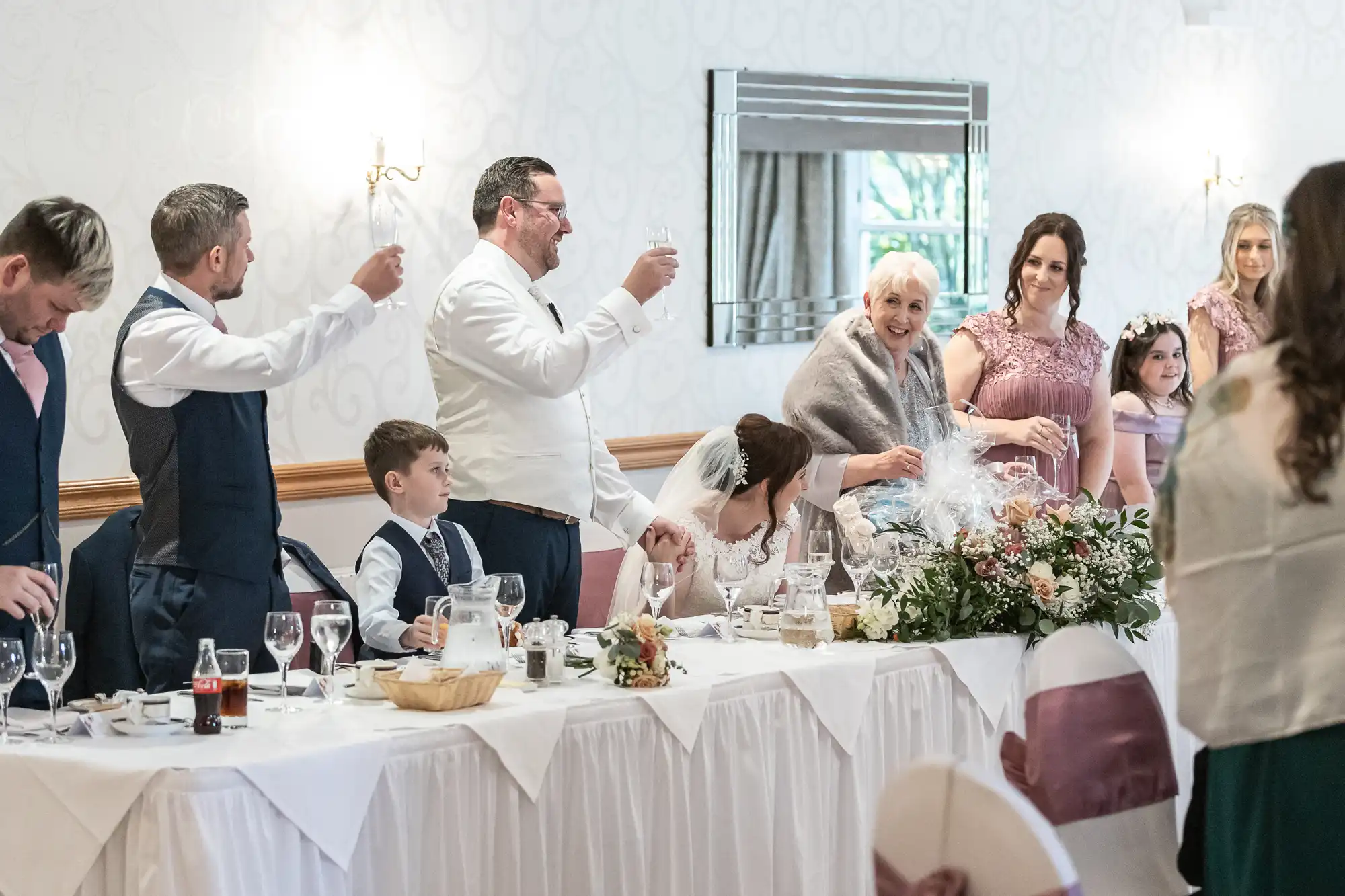 A wedding party sits and stands at a long, white-clothed table. The groom and others raise glasses in a toast, while the bride sits, smiling. Flower arrangements and tableware decorate the table. Guests of various ages are dressed formally, celebrating together.