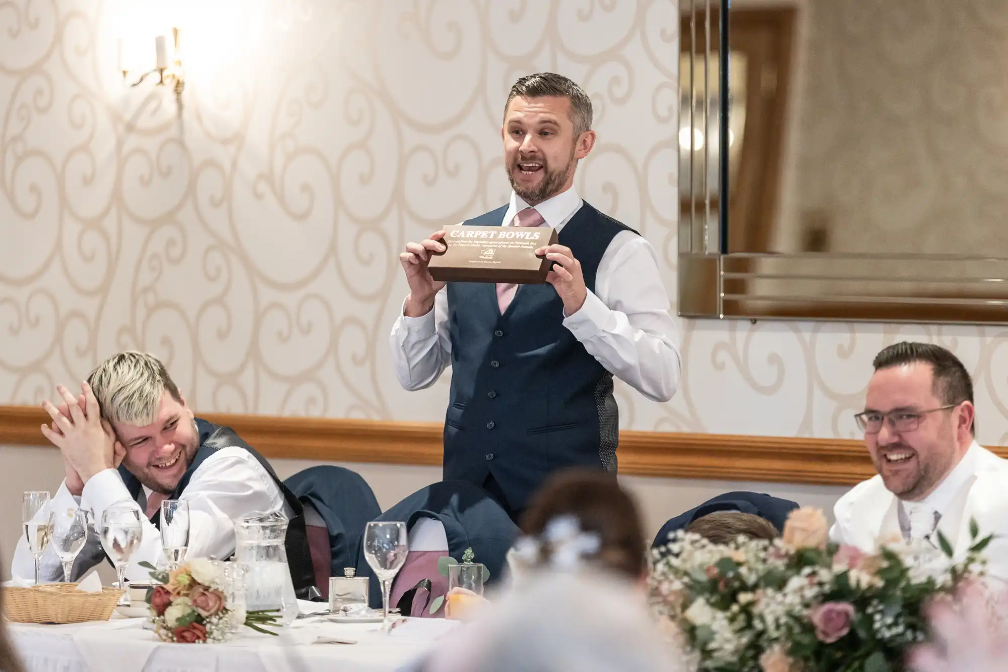 A man in a waistcoat stands at a wedding reception table, smiling and holding up a box labelled “Carpet Power.” Two men in suits seated nearby laugh, while the table is decorated with flowers, drinks, and crockery. The setting is bright with elegant wallpaper.