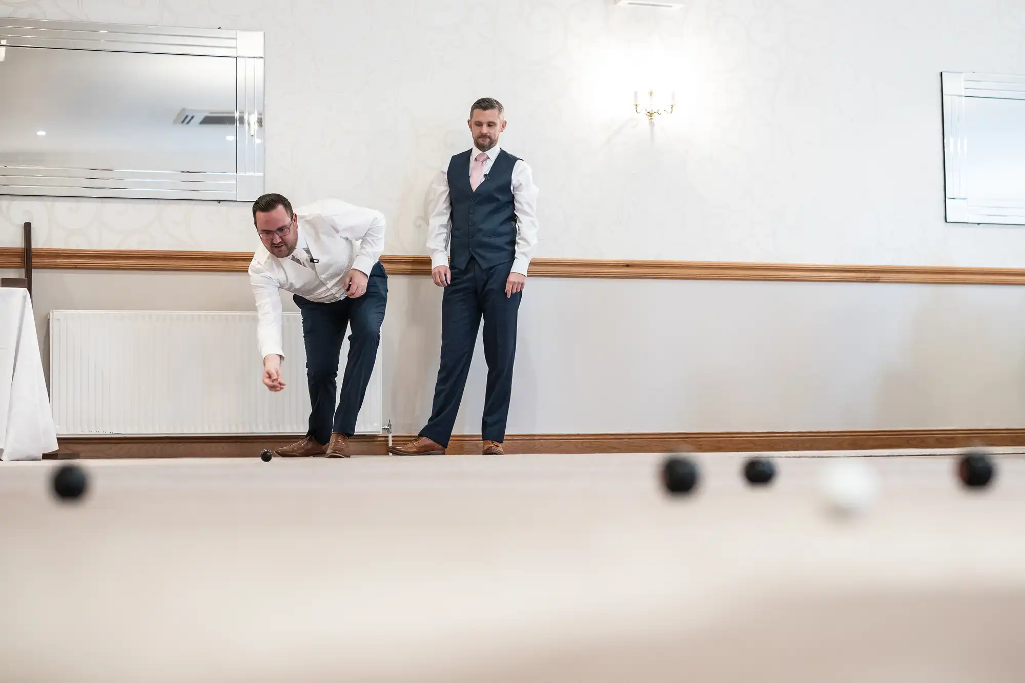 Two men in formal wear play an indoor game with black and white balls. One man bends down to roll a ball whilst the other stands nearby watching. The room has light walls, a wooden trim, mirrors, and a radiator in the background.
