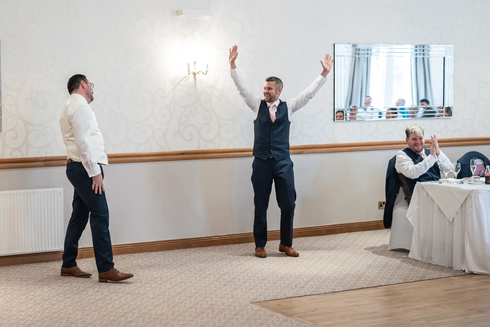 Two men in suits stand beside a white wall, one with arms raised in excitement. At a table to the right, a person in a white shirt and waistcoat claps while seated. The setting appears to be a formal event, likely a wedding reception.