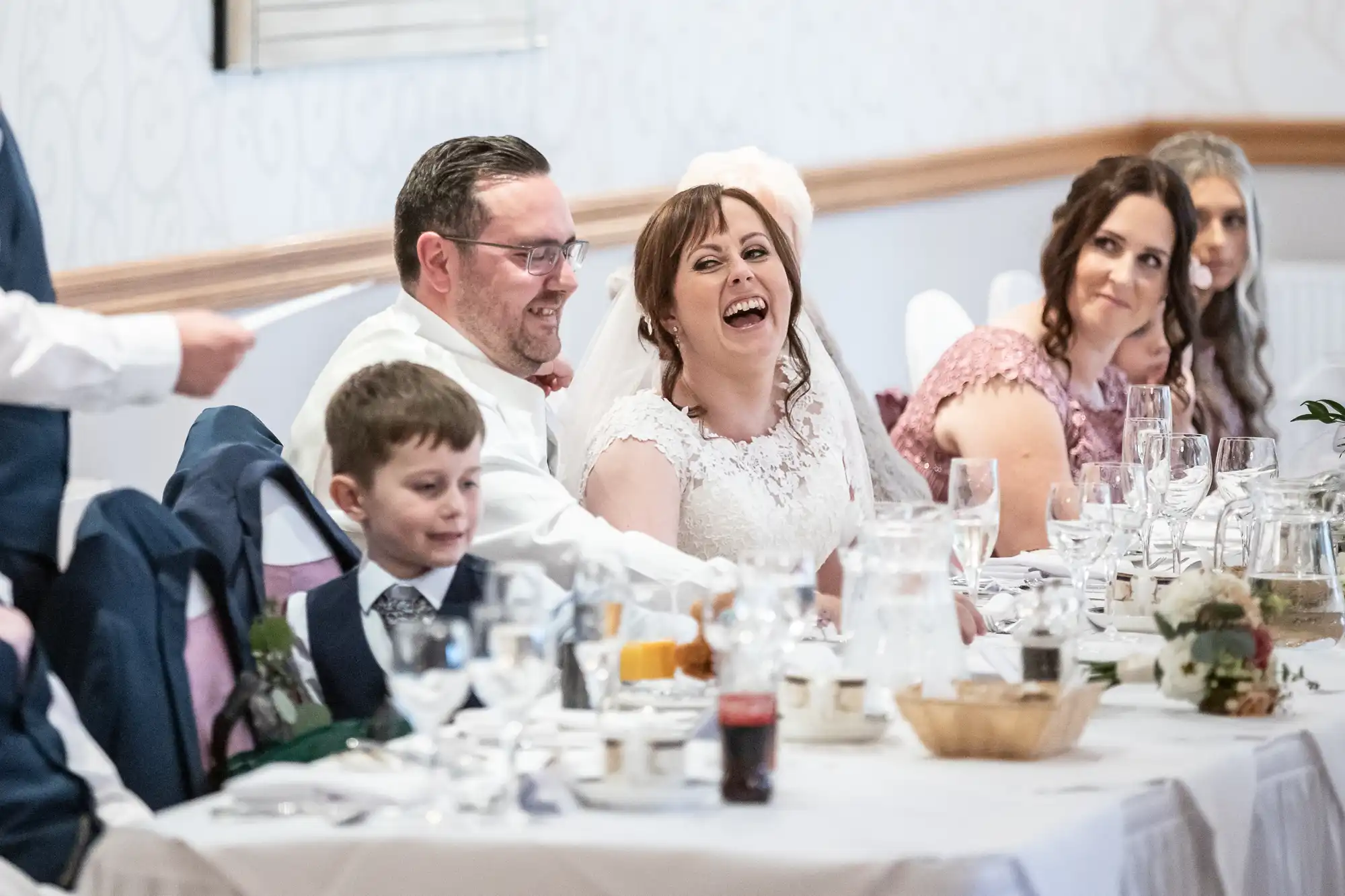 A bride and groom sit at a wedding reception table, laughing joyfully. A young boy in a suit sits nearby, and other guests, including women in formal dresses, are smiling. The table is set with glasses, plates, and flowers in a bright, elegant room.