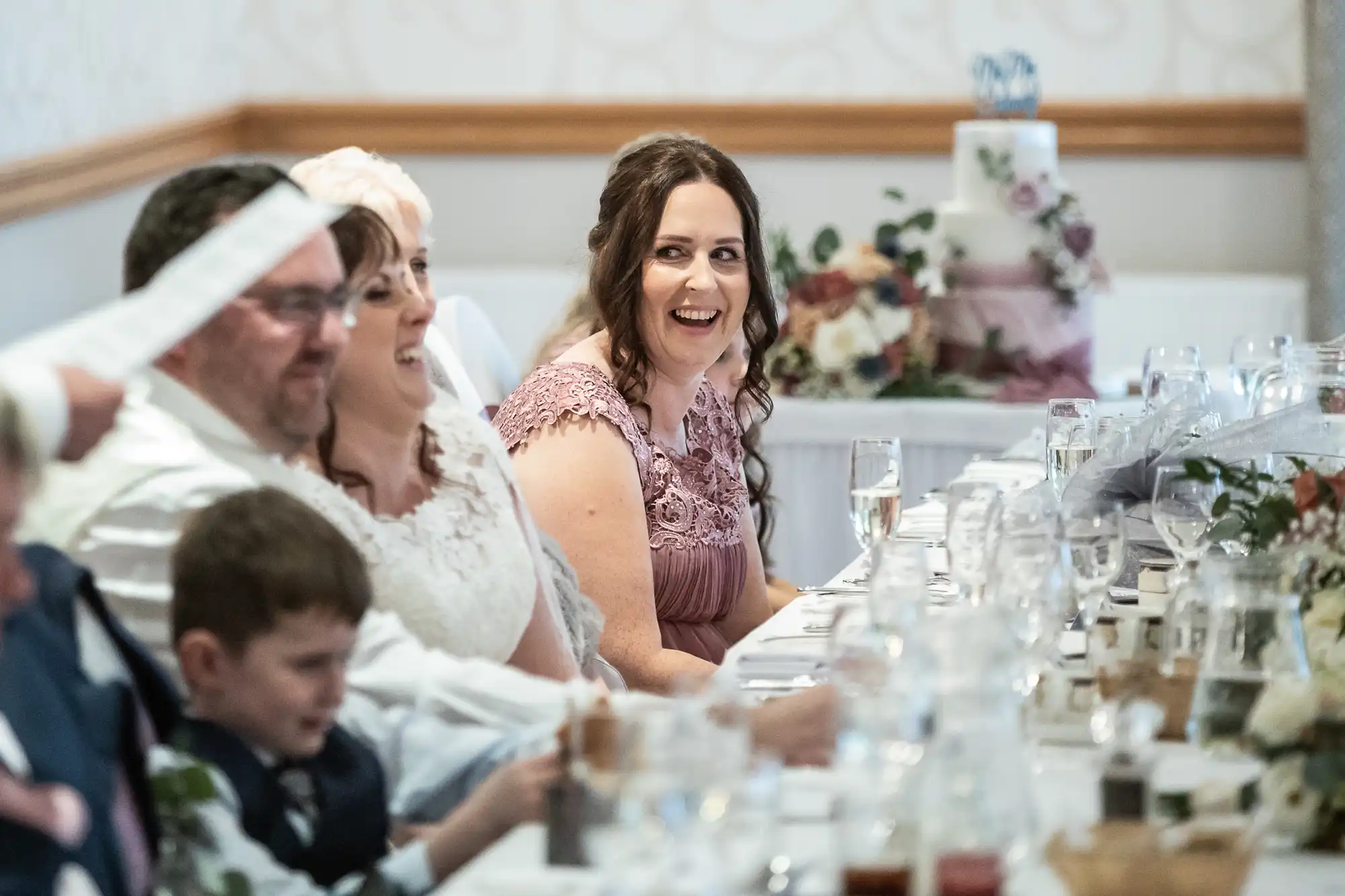 A group of people sit at a long wedding reception table, smiling and laughing. A woman in a mauve dress and a bride in white are at the centre. Glasses, flowers, and a tiered wedding cake are visible on the table and in the background.