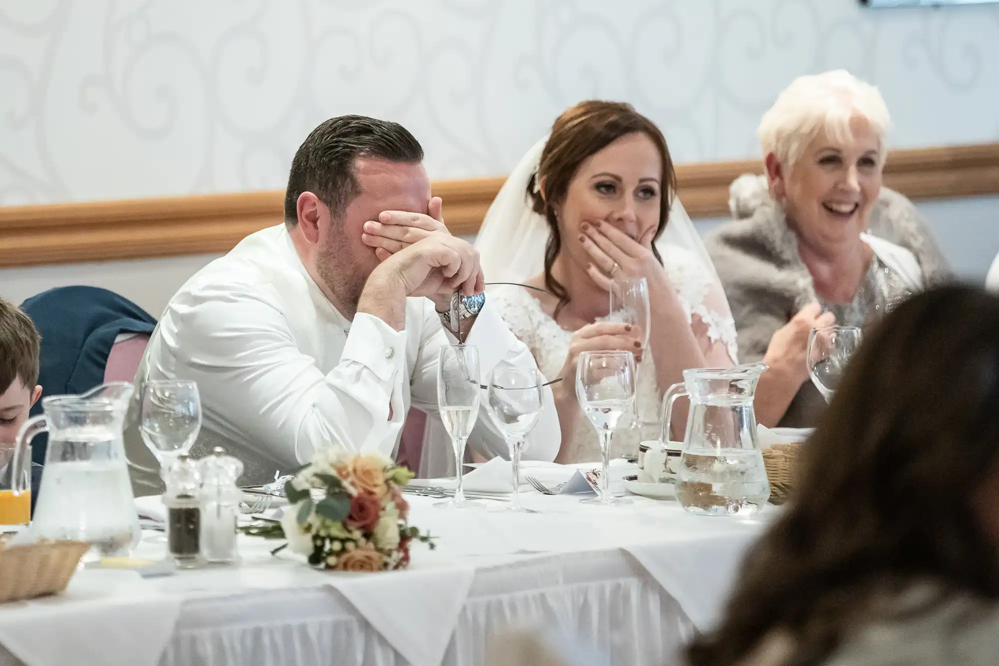 At a wedding reception, a groom in a white shirt covers his face, whilst the bride beside him in a veil smiles with her hand over her mouth. An older woman in grey sits next to them, laughing. The table is set with glasses, water jugs, and flowers.