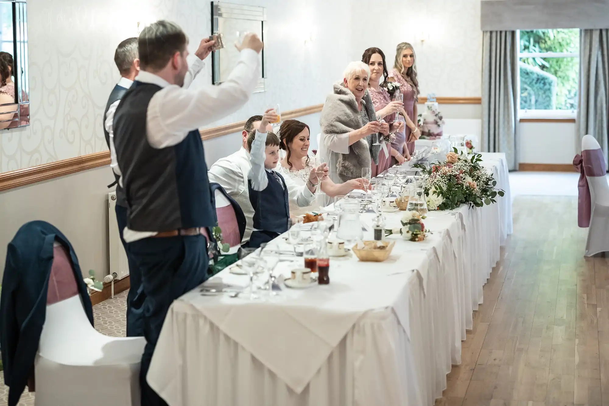 A wedding party stands at a long banquet table, raising glasses in a toast. The table is covered with a white tablecloth and set with plates, glasses, and flowers. People smile and celebrate in a bright, elegant room with large windows and soft lighting.