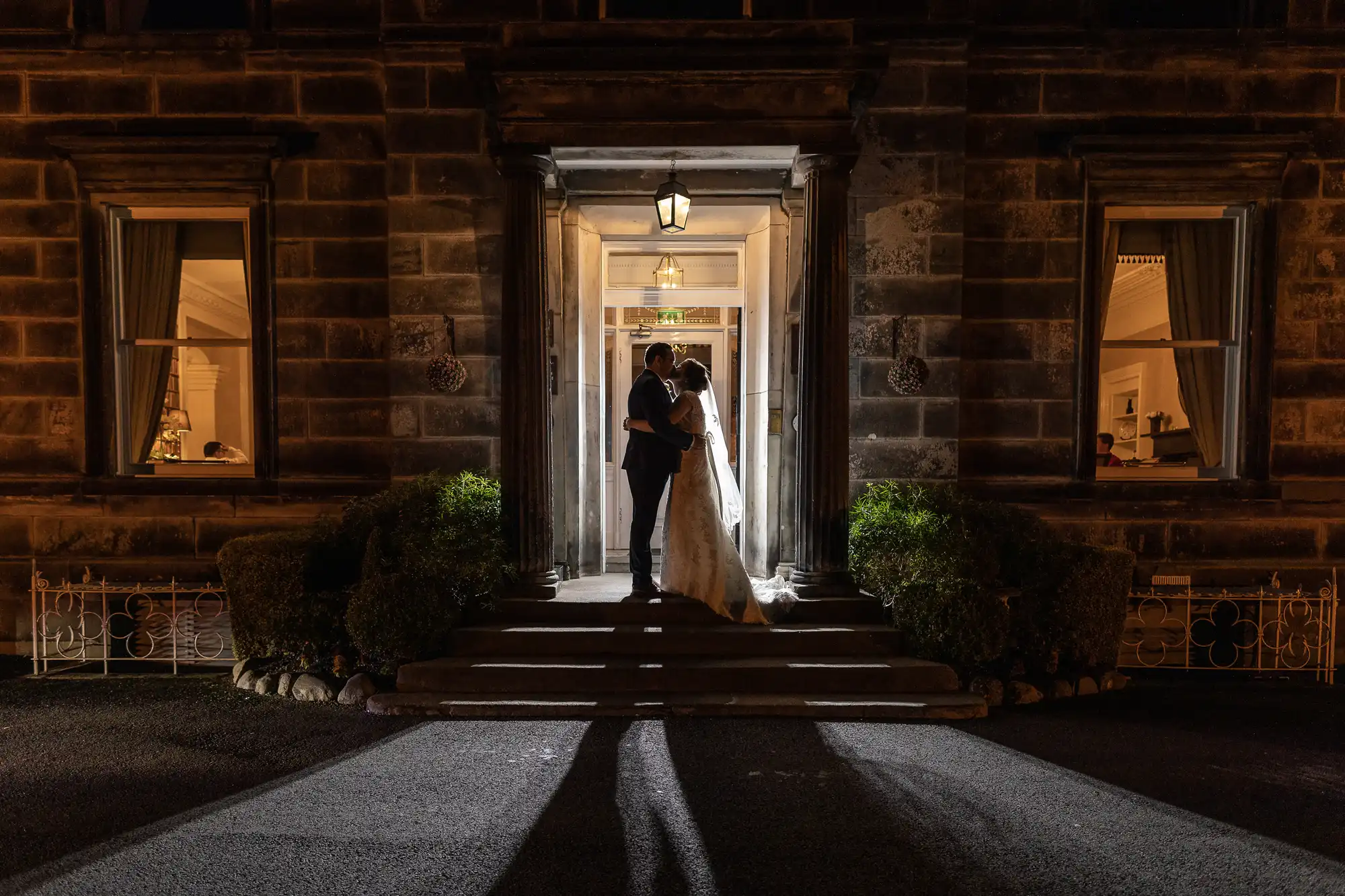 Garvock House Hotel wedding photographer image of a bride and groom stand in an embrace under a lit doorway of a stone building at night, casting long shadows behind them. The entrance is framed by two windows with curtains, and bushes line the steps leading to the door.